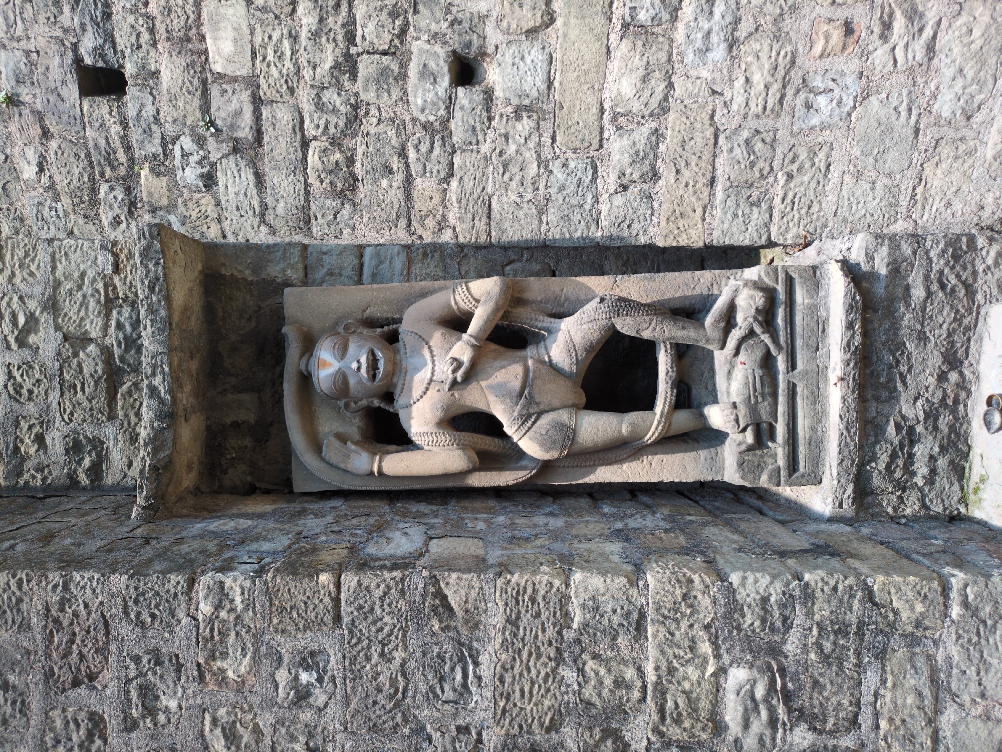 The image shows a stone carving of a deity set within a niche in a stone wall. The carving depicts a figure with a stylized face, adorned with jewelry and a loincloth. The figure is in a dynamic pose, with one leg bent and the other extended. A rope-like element is wrapped around the figure's legs. The surrounding wall is constructed from roughly hewn stone blocks, some of which appear weathered and cracked. The niche itself is rectangular, with a stone slab forming its base. The overall scene suggests an ancient or historical religious site. This carving is located in the Rewalsar area of Mandi, India.