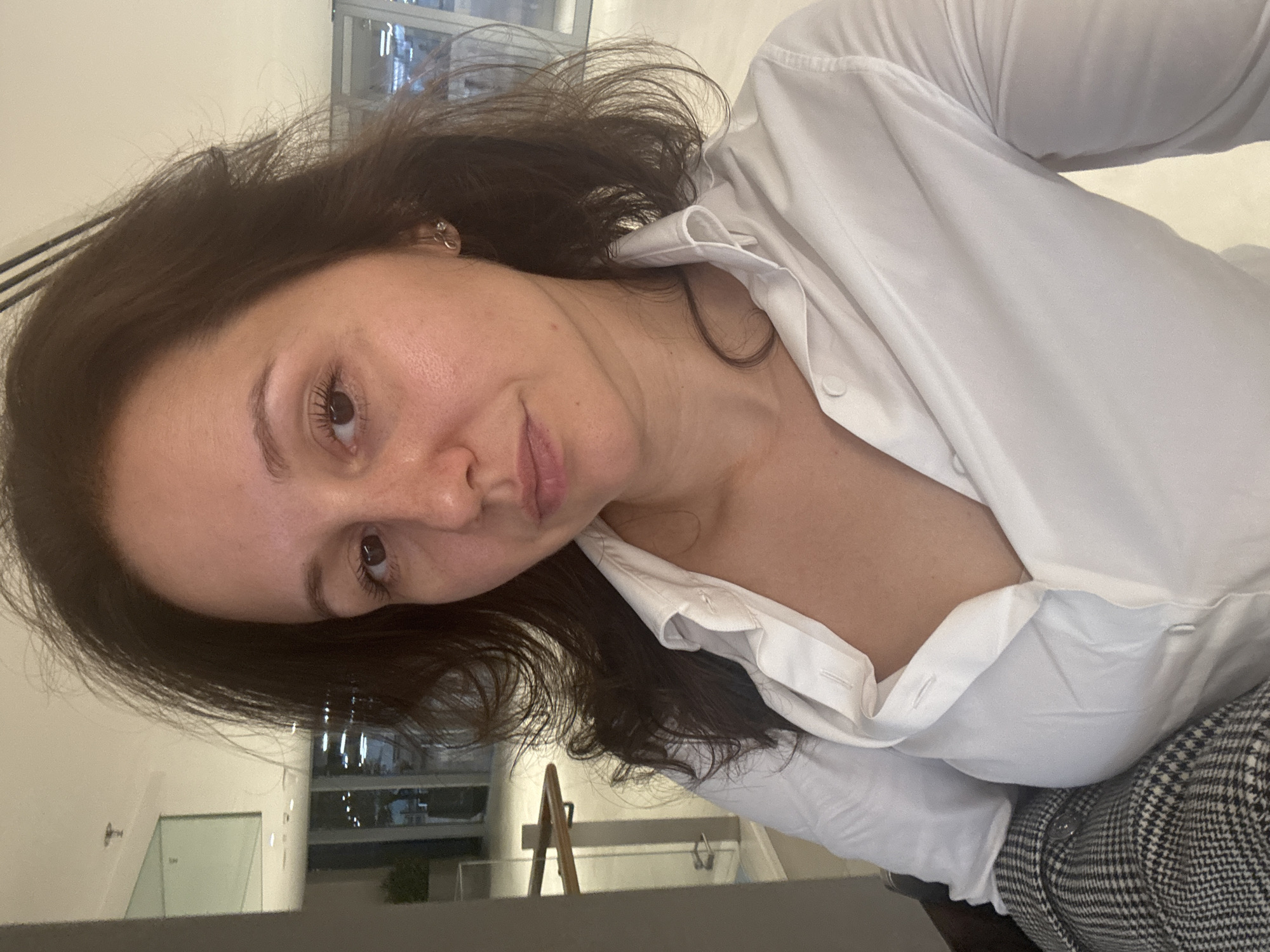 A woman with brown hair and wearing a white collared shirt and a grey and black plaid blazer is seated. She appears to be indoors in an office environment. The location is the city of Dubai, United Arab Emirates, as a building with a high-rise structure can be seen through a window in the background, consistent with Dubai's urban landscape.