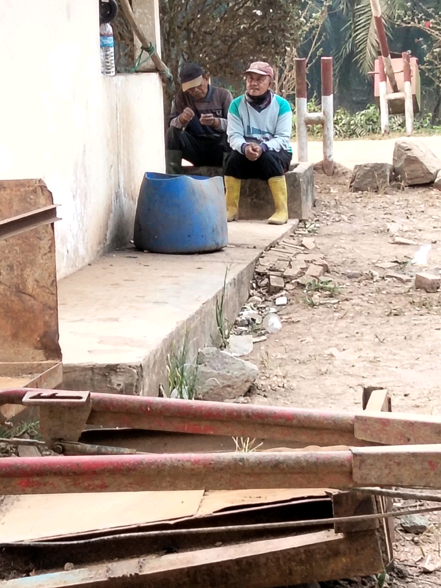 In Cinta Manis Lama, Indonesia, two men are seated outdoors near a building. The man on the left, wearing a dark long-sleeved shirt with stripes on the sleeves and dark pants, is looking down at his hands, which appear to be holding a small object. He is also wearing dark boots. To his right, a man wearing a light blue and teal long-sleeved shirt, black pants, a red baseball cap, and bright yellow boots is seated facing forward. He is looking to his left. Between the two men is a large, blue plastic barrel. The ground is a mix of dirt and scattered debris, including small rocks and pieces of brick. In the foreground, several rusty metal beams are visible, some with red paint. The background shows foliage and a wooden structure with red and white markings.