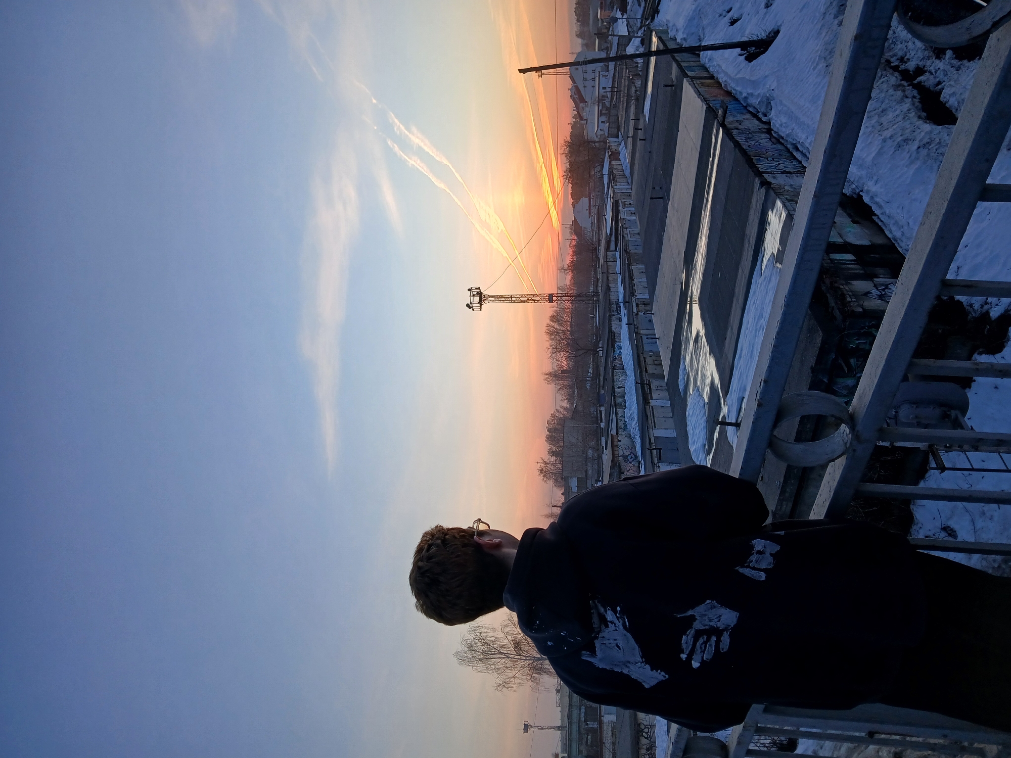 A person wearing a black hoodie stands on a concrete platform with metal railings, gazing at a sunrise or sunset in the distance. The sky transitions from blue to orange near the horizon. Contrails from aircraft are visible in the sky. Below, there is a partially snow-covered industrial area with rows of structures and a lattice tower. The location appears to be in an urban area of Chaikovsky, Russia.