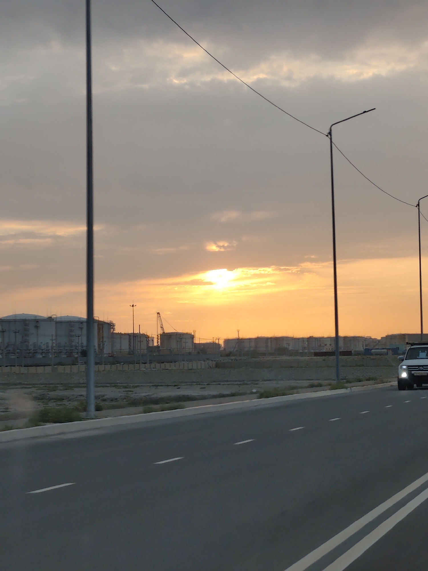 The image shows a highway in the city of Ömirzaq, Kazakhstan, at sunset. The sky is filled with dramatic clouds, illuminated by the orange glow of the setting sun. In the distance, industrial buildings and large storage tanks are visible. A white SUV is driving on the highway, its headlights on. There are several streetlights along the roadside, and power lines crisscross the sky. The road itself is asphalt with white lane markings. The overall scene depicts a vast, industrial landscape at the end of the day.