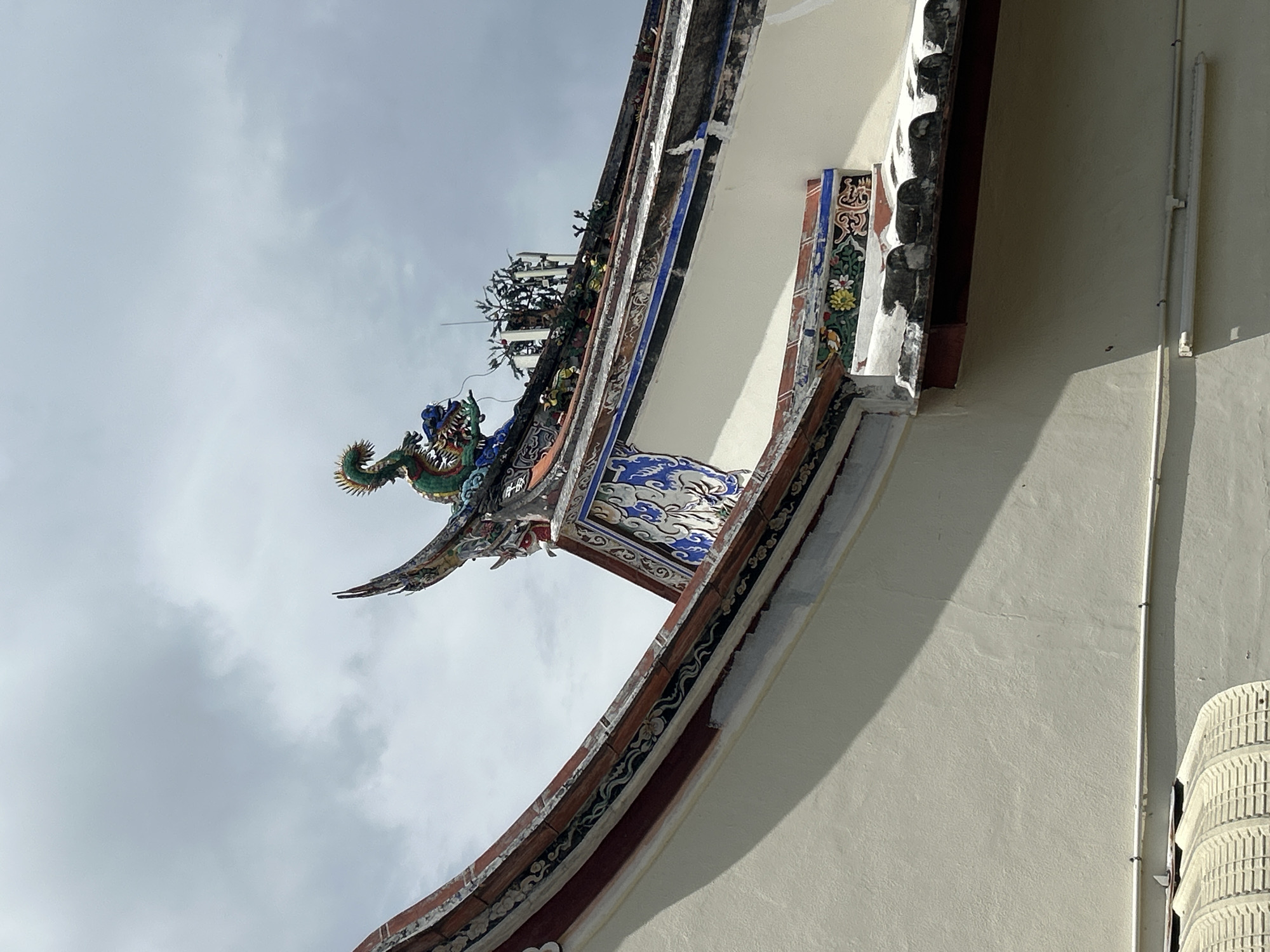 This is a medium shot, taken outdoors, of the roof of a traditional building in George Town, Malaysia. The sky above is overcast with light gray clouds. The building's roof features intricate architectural details, including curved eaves decorated with ceramic tiles and a prominent ceramic dragon sculpture at the peak of one of the gables. The dragon is green with red and blue accents. Other decorative elements on the roof include colorful tiled patterns and small, turtle-like figures. The wall of the building is painted a light cream color, and a section of a corrugated metal awning is visible at the bottom left of the frame. The overall impression is one of traditional East Asian architectural style.