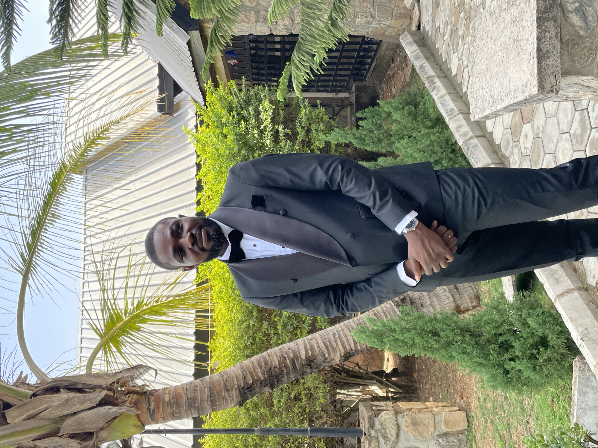 A man in a black tuxedo with a bow tie stands near a palm tree in an outdoor setting in Bukuru, Nigeria. He is wearing a wristwatch and has his hands clasped. The location includes greenery, stone steps, and a building with a corrugated metal roof.