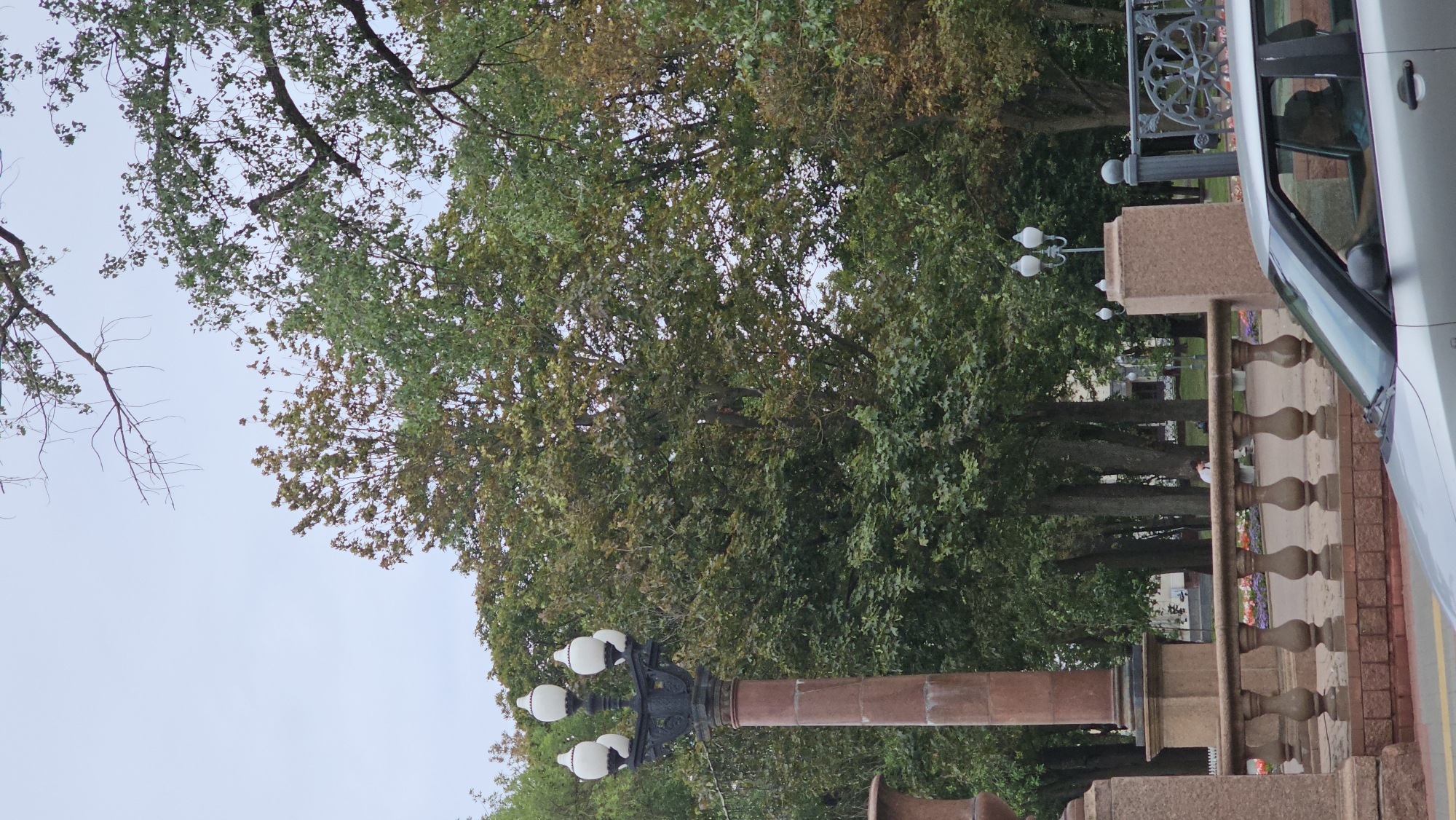 A vertical, medium shot captures an outdoor scene in a city park setting in Minsk, Belarus. In the foreground, the hood and side mirror of a silver car are visible, with a person driving. A stone balustrade runs horizontally across the middle ground, featuring decorative pillars. Behind the balustrade, trees with green and some autumnal foliage fill the background. To the left, a tall, ornate lamppost with four globes stands prominently on a stone pedestal. In the distance, more trees and some low-rise buildings are discernible. The sky is overcast.