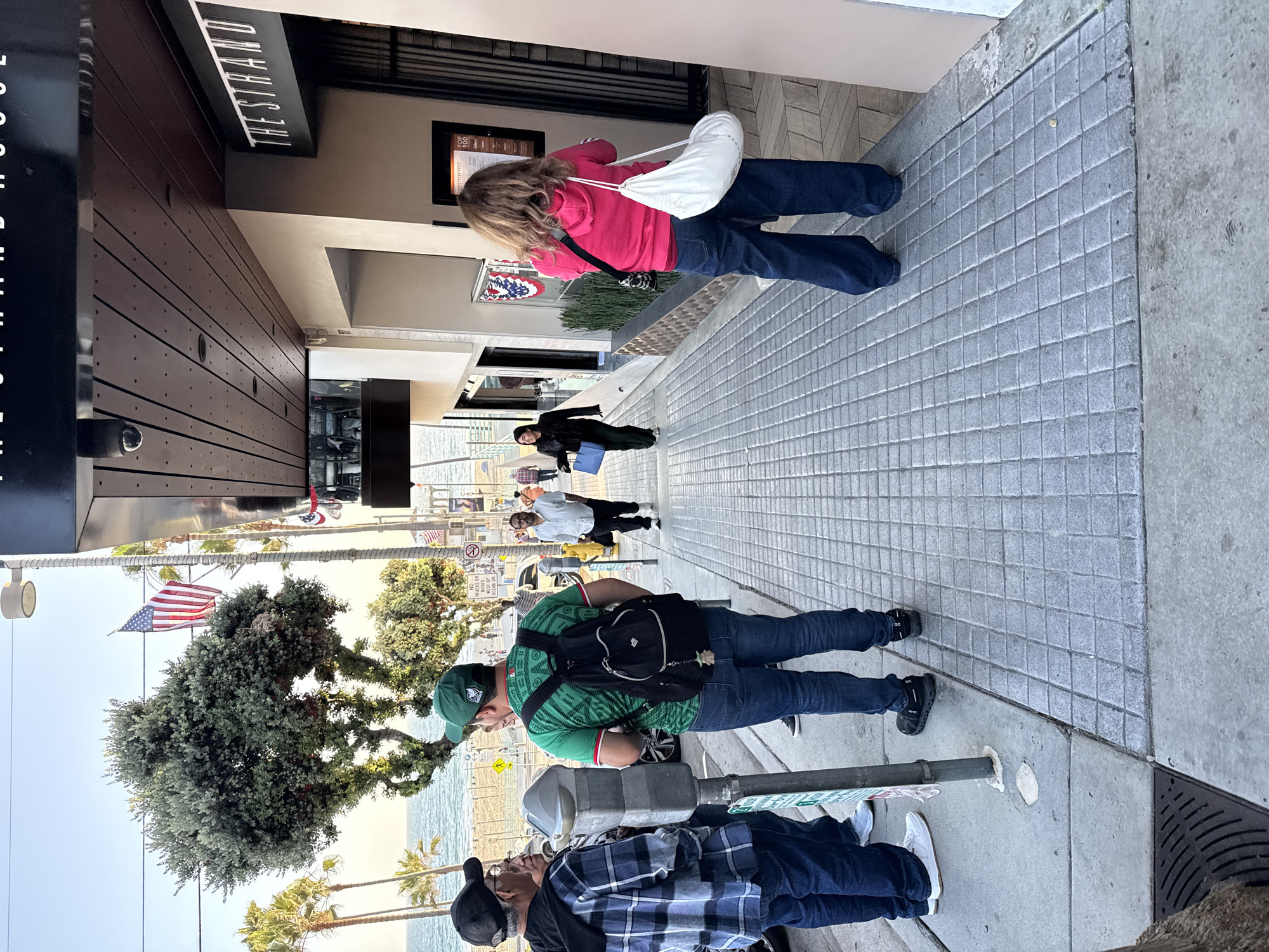 In Manhattan Beach, California, near the Strand, a group of people stand on a tiled sidewalk in front of a building that reads "THE STRAND HOUSE". An American flag waves from a pole. To the left of the frame, three men are gathered near a parking meter. The man closest to the viewer wears a blue and white checkered shirt and a black cap. Next to him, a man in a green shirt with a Mexican flag design and a green cap is looking away. The third man, partially visible, also appears to be male. Further down the sidewalk, a man in a white shirt and dark pants walks away from the camera. A woman in a black jacket and dark pants walks towards the camera carrying a blue bag. Another woman, wearing a bright pink hoodie and dark jeans, faces away from the camera, holding a white bag. Palm trees and the ocean are visible in the background.