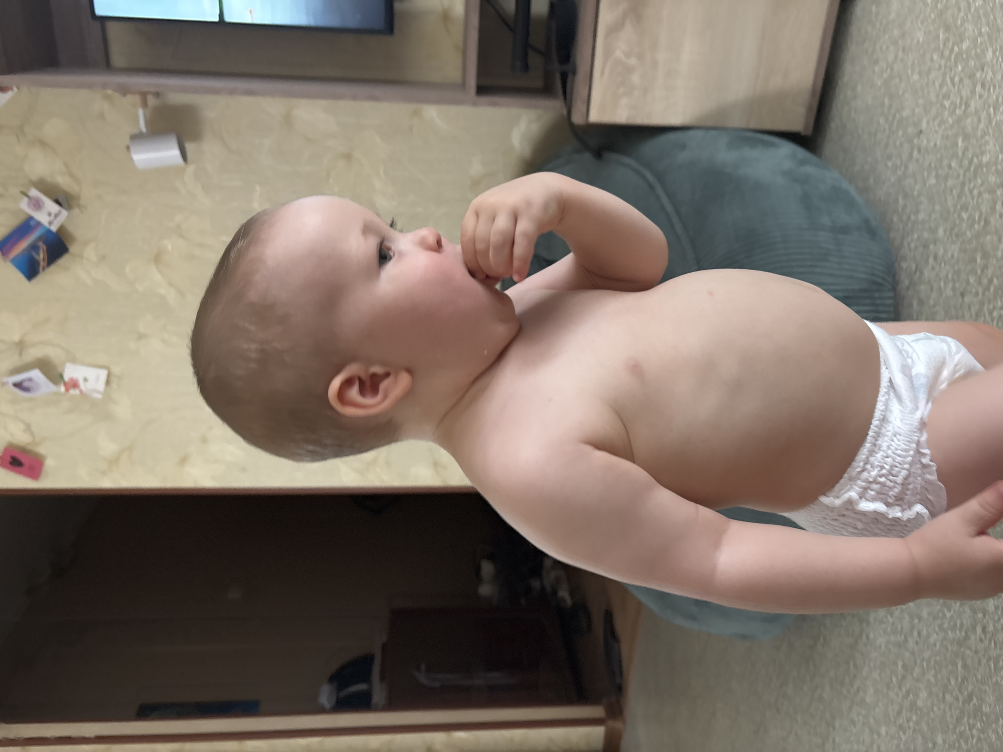 A young child, wearing a diaper, stands in a room and puts their hand to their mouth. The child has light-colored hair and is looking to the right. The background consists of a wall with what appears to be wallpaper, a dark doorway, a television on a stand, and a large, dark green beanbag chair. The floor is covered in a light-colored carpet. The image appears to be taken indoors in a residential setting. There is no information available to determine the location in the city of Naberezhnye Chelny, Russia.