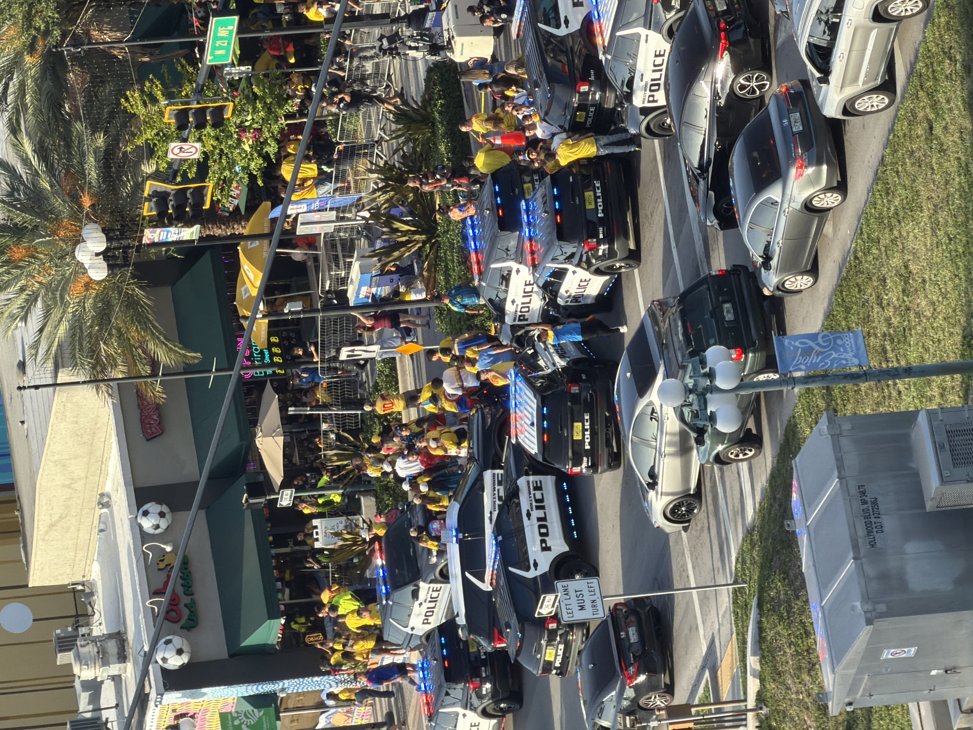 The image captures a busy street scene in Hollywood, Florida, USA, likely during an event or gathering. Multiple police vehicles, specifically marked "HOLLYWOOD POLICE," are present, with their lights illuminated, suggesting an official presence or a response to a situation. A crowd of people, many wearing yellow and red shirts, are gathered on the sidewalks and in the street, some observing the police activity and others appearing to be part of a procession or celebration. The environment includes buildings with storefronts, palm trees, and traffic signals, indicative of a commercial and urban area. One building has a sign that appears to read "Oli's Mexican Restaurant," and there are soccer balls decorating its exterior. A traffic sign indicates "N 21 AVE." The overall atmosphere seems to be one of crowd management and public order.