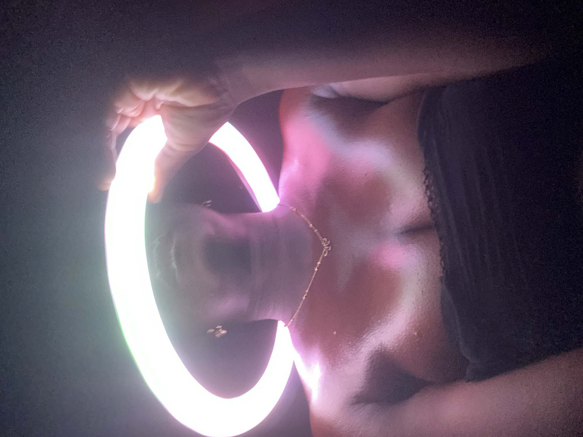 A person is holding a ring light in front of their face and chest, with the light illuminating their skin. A delicate gold necklace is visible around their neck. The person has dark hair and is wearing a black top. The background is dark. The location of Maigizo, Nigeria, is not discernible from the image.