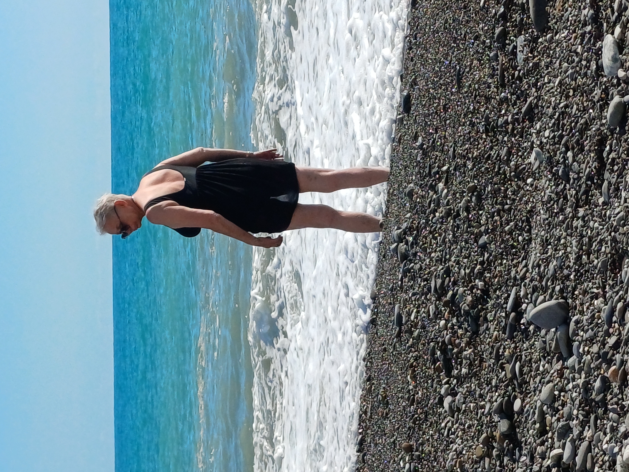 An elderly woman with short, grey hair and sunglasses is standing in the shallow surf on a pebble beach. She is wearing a black one-piece bathing suit and is facing away from the camera, looking out at the turquoise ocean. The waves are creating white foam as they crash on the shore. The sky is a clear blue. The beach is covered in dark grey and brown pebbles. The location is on the coast of Sochi, Russia.