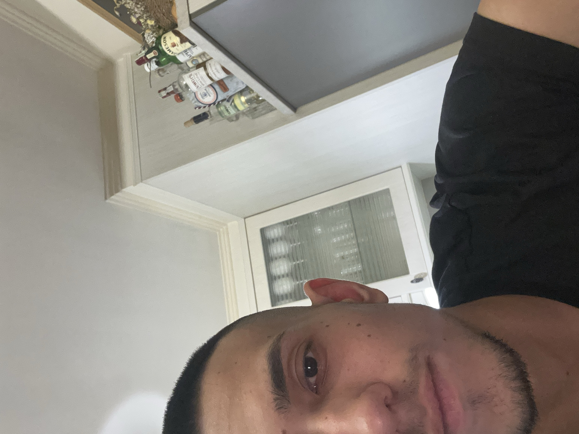 The image depicts a partial view of a young adult male in an indoor domestic environment, situated in Plovdiv, Bulgaria. The subject presents with short, dark, cropped hair, a visible right eye, a prominent left ear, and sparse facial hair comprising a mustache and goatee. He is clad in a black t-shirt. The background on the left reveals a white cabinet equipped with glass doors, housing multiple clear drinking glasses. To the right, a white wall exhibits decorative crown molding near the ceiling. A shelf above the subject's right shoulder supports an arrangement of liquor bottles, specifically identifiable as Jameson Irish Whiskey, Dewar's White Label Scotch Whisky, and Bombay Sapphire Gin, alongside an indistinct dried botanical or woven decorative object.