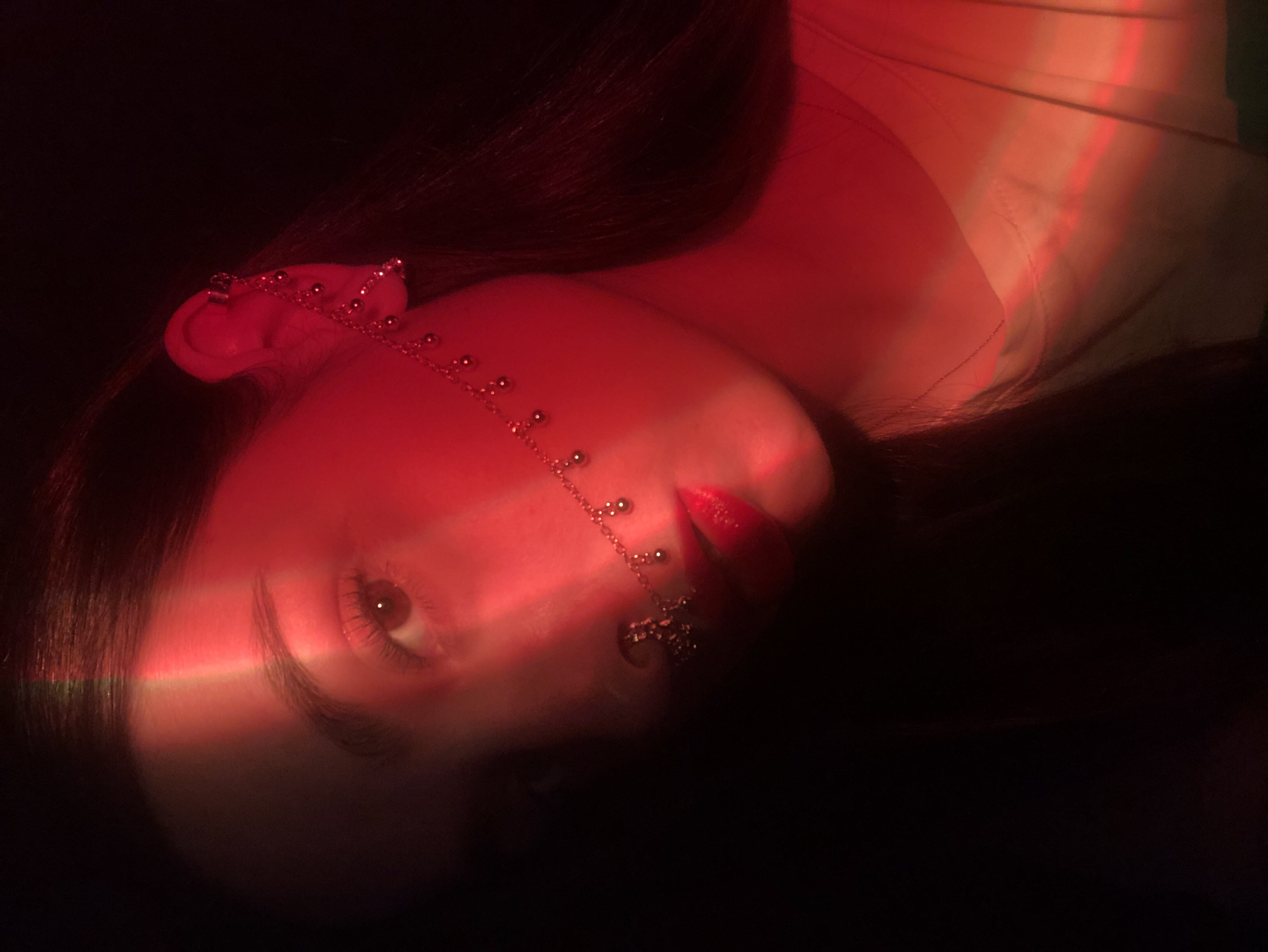 A close-up, rotated, and obscured by a red light photograph shows a person's face, turned sideways and upward. The person has fair skin, dark hair, and a piece of jewelry attached to their ear and running along their face. The person's eye is open. Their lips are red. The background is dark. The location in the city of Noviy, Russia, is unknown.