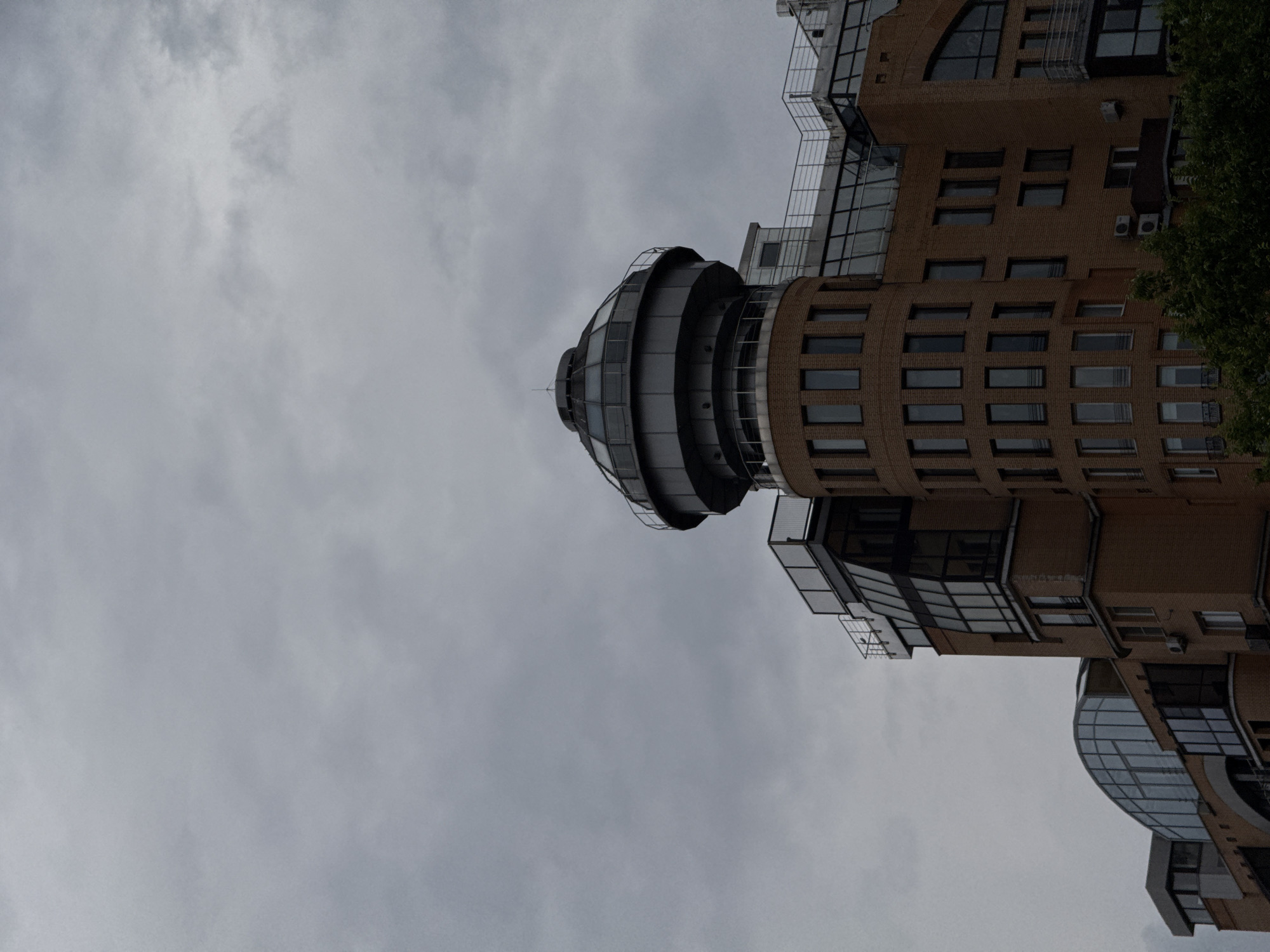 The image is an upward shot of a modern, multi-story building with a distinctive rounded, glass-enclosed top section. The building's facade is a mix of light brown brickwork and dark-tinted windows, arranged in horizontal bands on the lower floors and in a more complex geometric pattern on the upper sections. There are balconies with metal railings and visible air conditioning units on some of the brick sections. A sliver of green foliage is visible on the far right of the image, suggesting the building is surrounded by trees. The sky is overcast and gray, indicating a cloudy day, possibly with impending rain. The orientation of the image is such that the top of the building is at the bottom of the frame. There is no discernible activity or people in the frame. The overall impression is one of urban architecture under a muted sky.