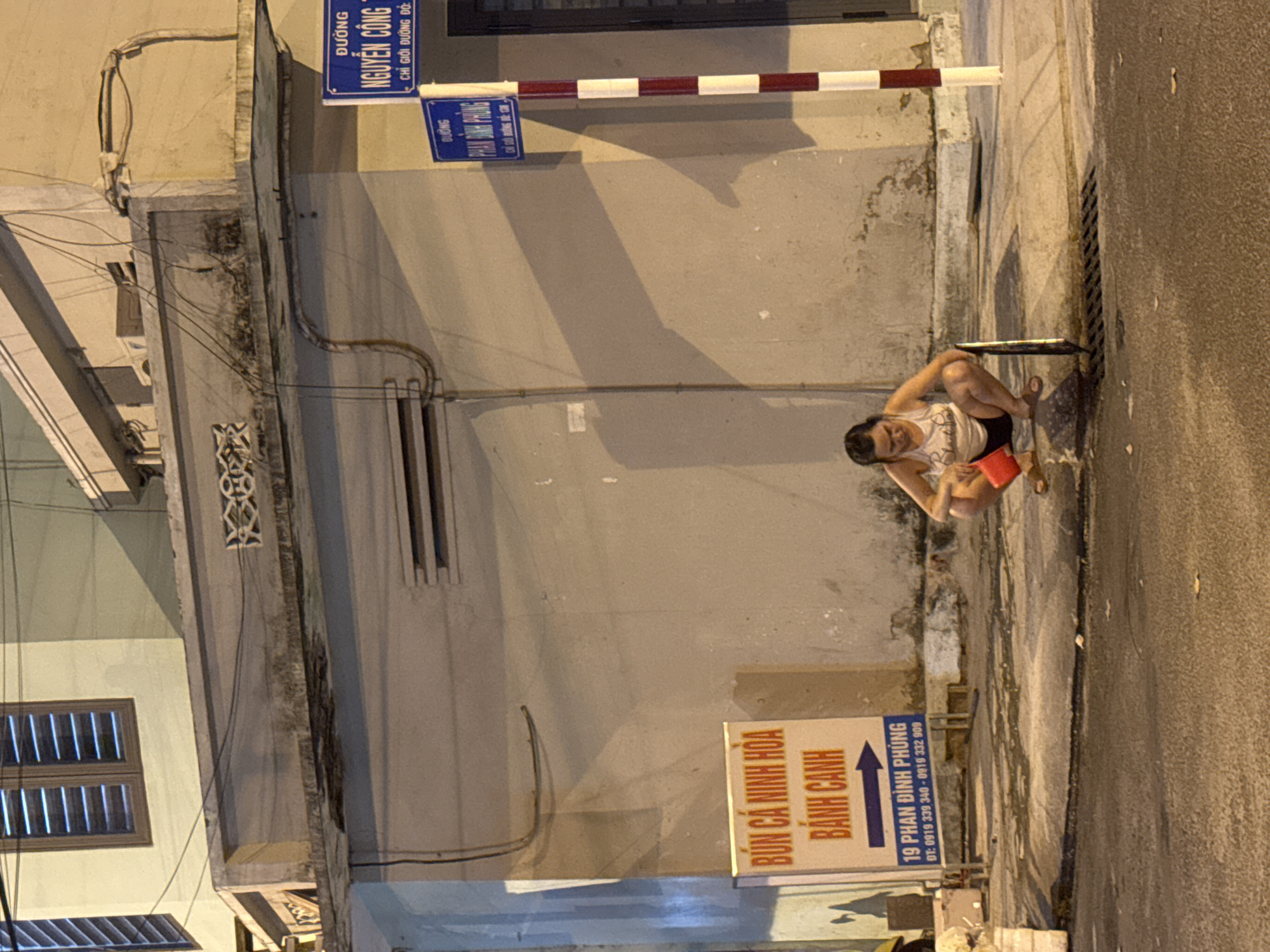 The image depicts a street scene at night in Nha Trang, Vietnam. In the foreground, a woman is squatting next to a drain. She is wearing a white tank top and dark shorts, and is holding a red bucket. A metal grate is visible on the drain. To the left of the woman, a sign with Vietnamese text advertising "BÚN CÁ NINH HÒA" and "BÁNH CANH" is mounted on a pole. The sign also includes a phone number and an address: "19 PHAN ĐÌNH PHÙNG". In the background, there is a building with a cream-colored wall and electrical wires running along its exterior. Two street signs are visible on a pole to the right, one reading "ĐƯỜNG NGUYỄN CÔNG TRỨ" and another, partially obscured, reading "ĐƯỜNG PHAN ĐÌNH PHÙNG". The street is paved with asphalt and there are scattered leaves on the ground. The lighting suggests it is dusk or nighttime.