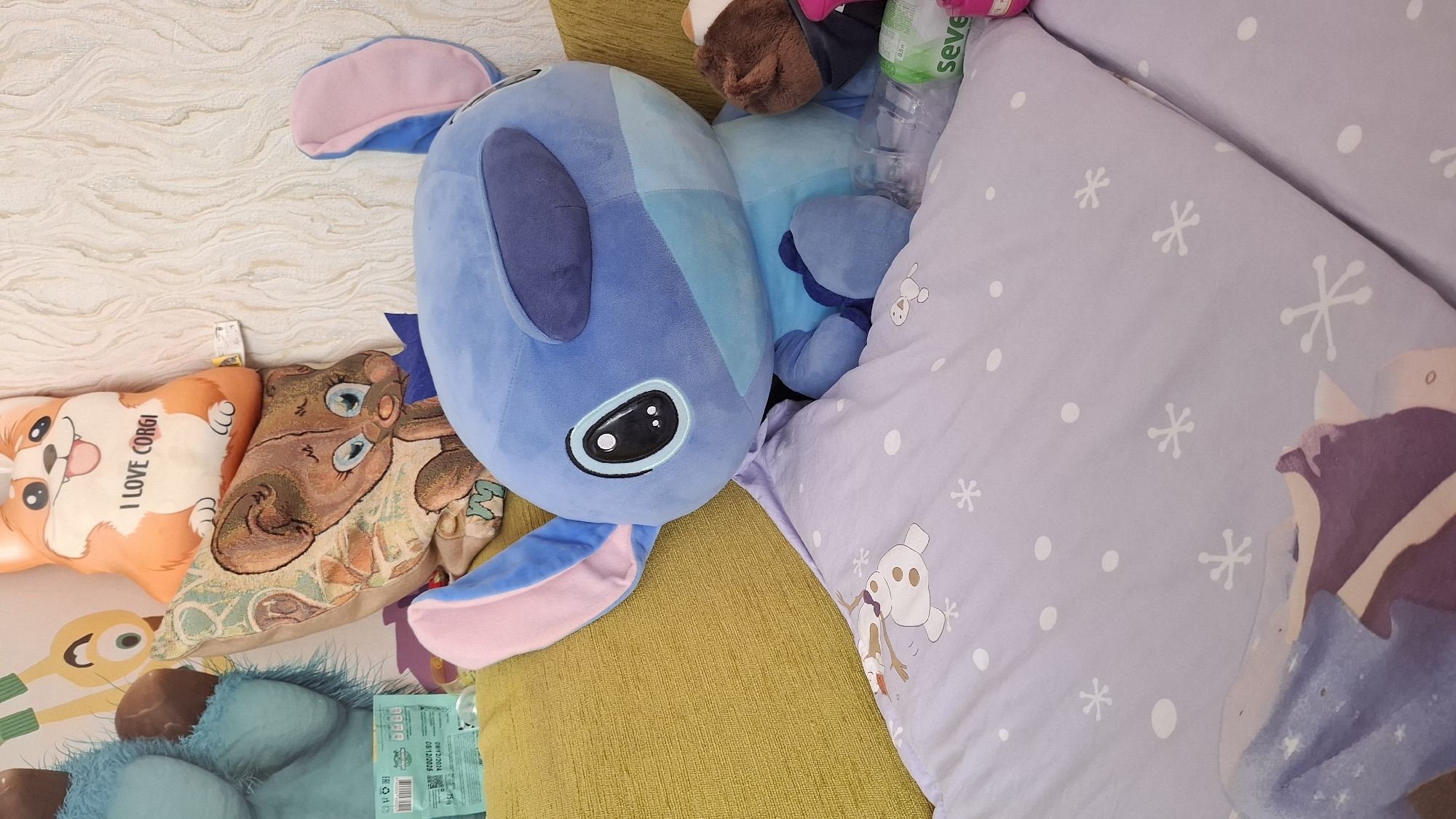 A stuffed toy resembling the character Stitch from Disney's Lilo & Stitch is the primary subject of the image, positioned on a couch with a yellow cushion. Stitch is large and blue, with characteristic big ears and eyes. It is draped over a light purple blanket that features a pattern of white snowflakes and cartoon characters, possibly from the movie Frozen. Behind Stitch, several other stuffed pillows are visible, including one with a corgi design that says "I LOVE CORGI" and another with a brown bear or mouse character. A portion of a turquoise plush object, possibly another stuffed toy, is visible on the left side of the frame. In the lower right corner, part of a plastic water bottle and a green and white package labeled "seve" are present. The background appears to be a textured wall with a light color. The image does not contain any visual information that allows for the identification of its location in Shymkent, Kazakhstan.