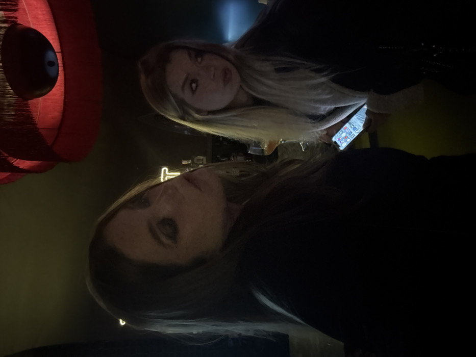 Two women pose in a dimly lit setting. The first woman, with dark hair, is situated in the foreground. Behind her, the second woman has blonde hair and is looking at her phone. A red, layered lamp is visible in the background on the left. The lighting is poor.