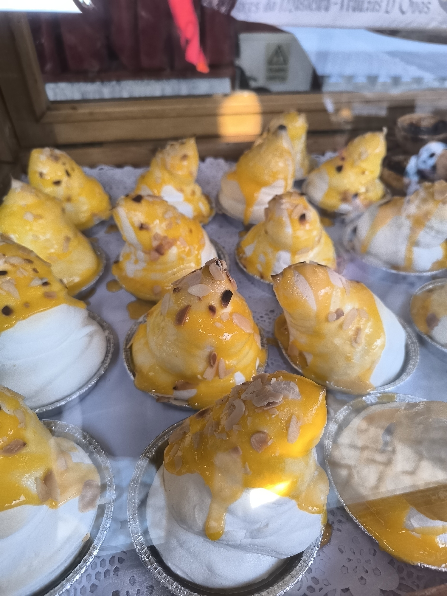 The image depicts a close-up view of a display case filled with several meringue-based desserts, likely sold in the historic town of Óbidos, Portugal. The desserts are characterized by their white meringue base, topped with a vibrant yellow sauce, possibly a fruit coulis or custard, and sprinkled with slivered almonds. The desserts are arranged in individual foil tartlet molds, some with decorative paper doilies underneath. In the background, a blurred sign suggests the location may be a confectionery or bakery, and the presence of what appears to be a price list or product information adds to this impression. The overall lighting suggests an indoor setting, possibly a market stall or a shop. There are no people visible in the image. The desserts are the sole focus, presented in an appealing arrangement.