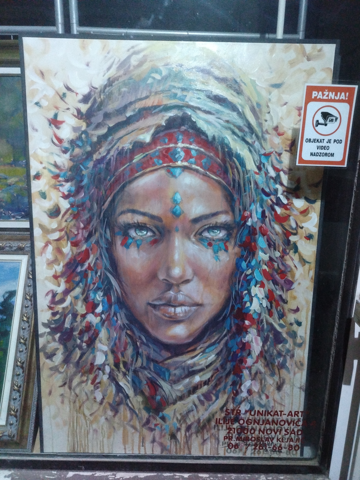 The image shows a painting of a woman with a headdress, framed and displayed. A sign reading "PAŽNJA! OBJEKAT JE POD VIDEO NADZOROM" (ATTENTION! THE OBJECT IS UNDER VIDEO SURVEILLANCE) is attached to the painting's frame. Below the painting, text indicating "STR 'UNIKAT-ART' ILIJE OGNJANOVIĆA 21000 NOVI SAD PR. MIROSLAV KEJAJ 067 / 261-66-80" is visible, identifying the art shop as "Unikat-Art" located at Ilije}}^{(\text{Ilija})}{}$ Okanovića street in Novi Sad, Serbia. The painting itself is colorful, featuring the woman's face with detailed eyes and a decorated headdress made of various colored brushstrokes resembling feathers or petals. The background is abstract and light-colored. The painting is leaning against another framed artwork on the left, and the overall scene appears to be within an indoor setting, possibly a gallery or shop.