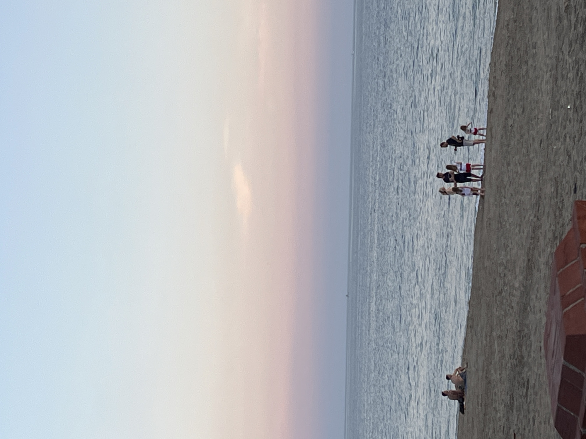 This image captures a serene beach scene, likely at sunset or sunrise, given the soft gradient of colors across the sky. The sky transitions from a pale blue on the left to a delicate pink and orange hue towards the right, meeting a calm, rippled body of water, presumably the Mediterranean Sea. The horizon line is clear and extends across the width of the image.
The foreground features a sandy beach, where several people are present. A group of five individuals, appearing to be a mix of adults and younger people, stands near the water's edge on the right side of the image. They are dressed in casual attire and seem to be observing the sea or interacting with each other. Further along the beach, towards the bottom right, two individuals are seen relaxing on the sand, one lying down and another sitting. Adjacent to them is a distinctive structure made of reddish-brown bricks, possibly a low wall or part of a building. In the far distance on the tranquil sea, a small, faint object, possibly a boat or buoy, is visible on the horizon.
The overall atmosphere conveys leisure and relaxation, typical of a coastal evening or early morning. The weather appears clear and pleasant. There is no visible text in the image. The setting is consistent with a scenic beach in Alanya, Turkey.