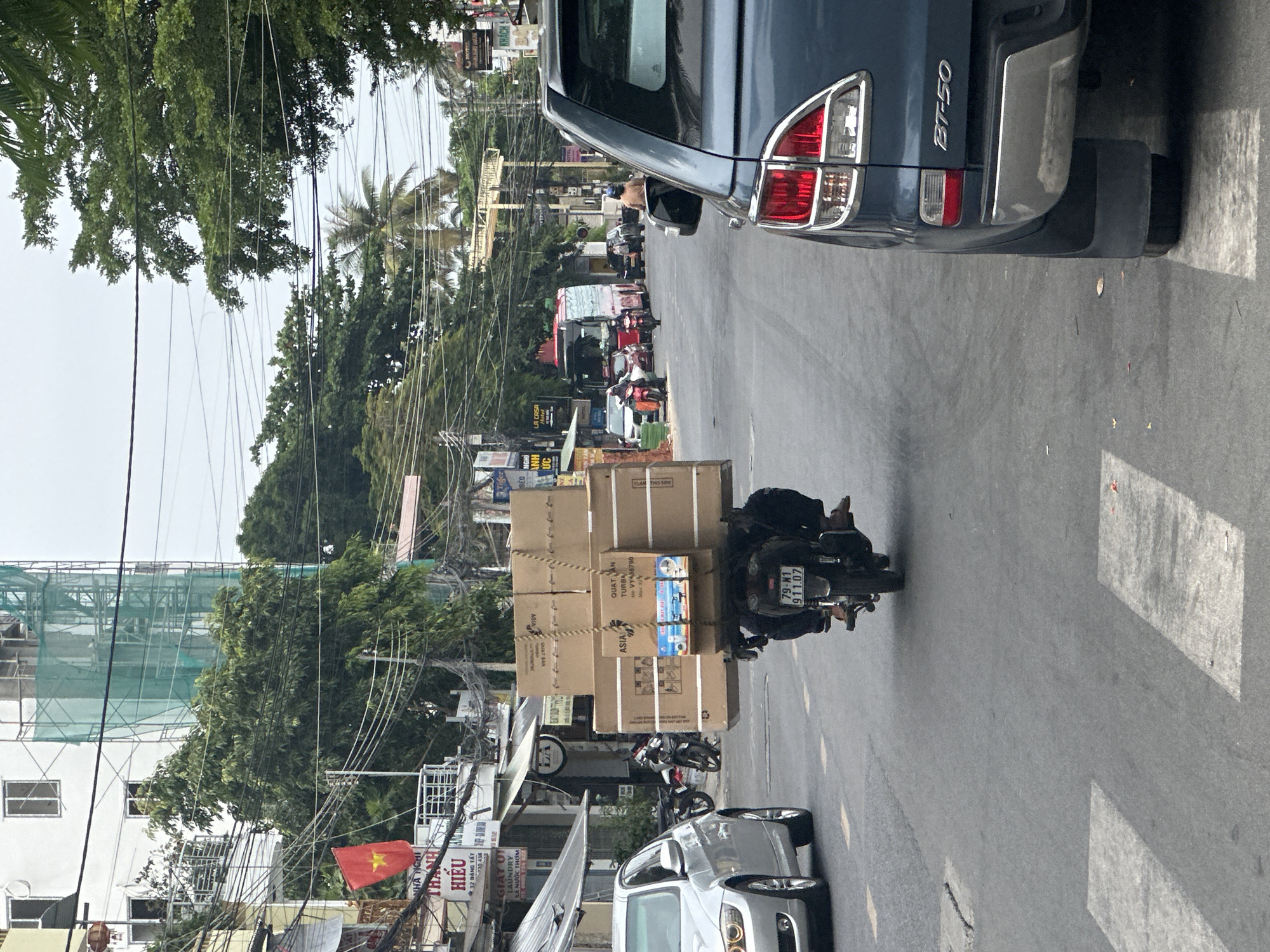 The image captures a busy street scene in Bac Nha Trang Ward, Vietnam. A motorcycle is heavily loaded with a stack of cardboard boxes secured with rope, moving away from the camera. To the right, a blue BT-50 pickup truck is parked. On the left, a silver car. There are buildings and street signs lining the side of the road, including a Vietnamese flag displayed outside a business named "Hieu Anh", which appears to be a laundry service and inn or motel. Power lines crisscross overhead amidst the trees. In the distance, there are other people and vehicles on the road. The motorcycle's license plate reads "79-N1 911.07". The boxes are labeled “ASIA” and “QUATAN TURBA”.