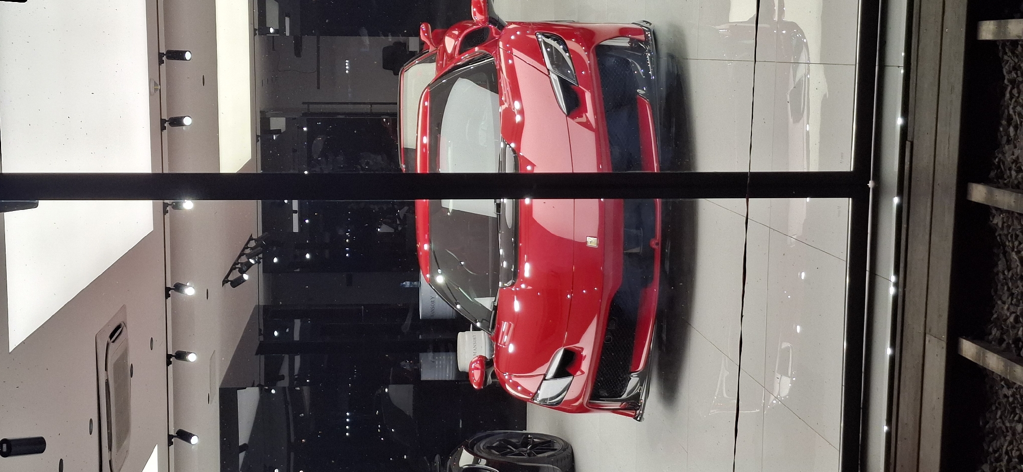 The image shows the front of a red Ferrari sports car parked inside a showroom. The car is the primary subject, with its sleek design and bright red color prominently displayed. The showroom appears to be well-lit, with spotlights on the ceiling and reflective flooring. Behind the Ferrari, another red car is partially visible, along with dark interior walls and displays. The scene is viewed through a large window, with a black vertical pillar obscuring a portion of the view. In the foreground, there are some wooden planks and gravel, suggesting the car is displayed behind a barrier or in a street-facing showroom. The presence of "Mansory" branding on cylindrical objects in the background indicates a connection to high-end automotive customization. The lighting suggests it is either evening or the showroom's artificial lighting is dominant.