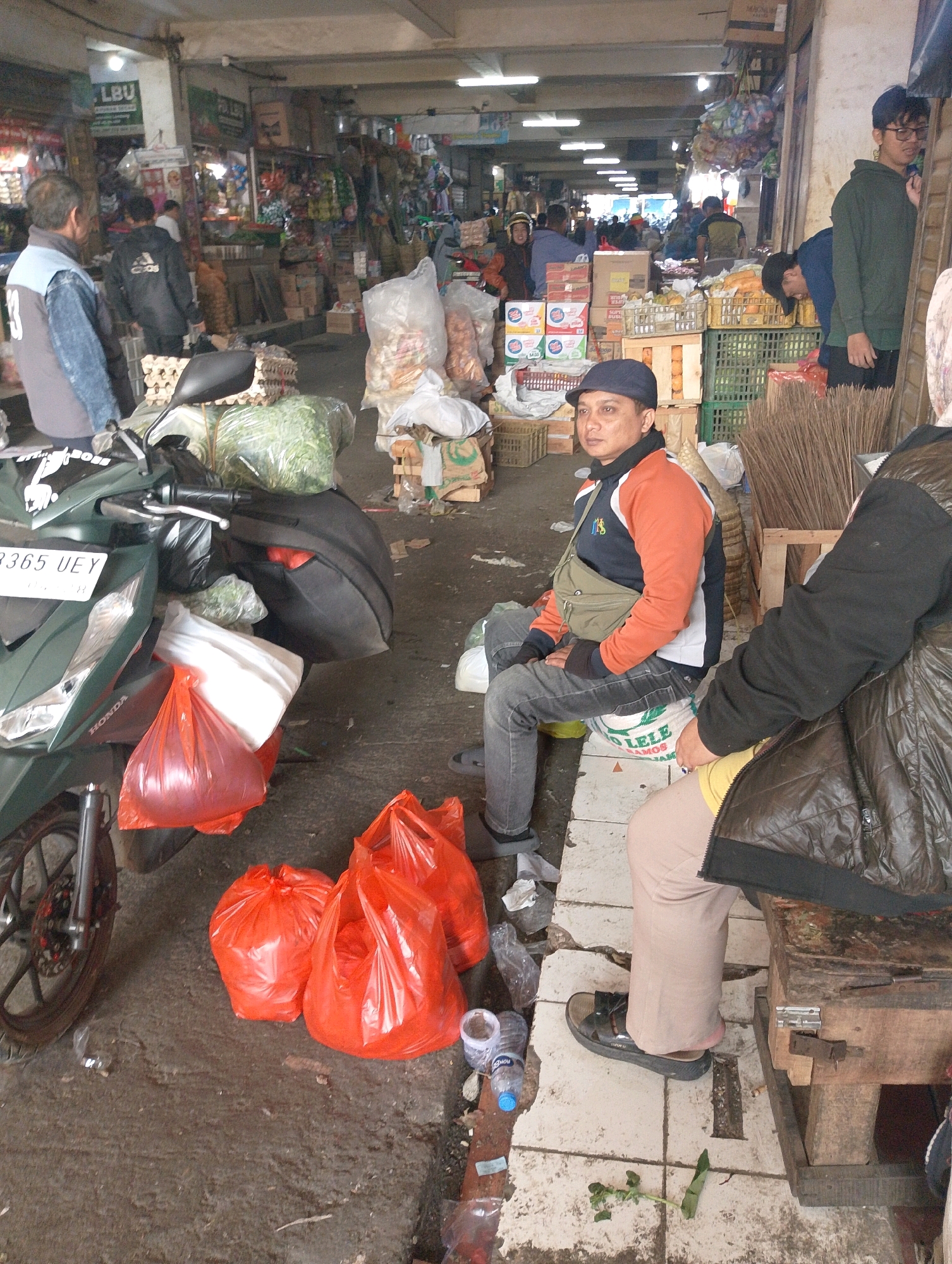 The image depicts a busy marketplace scene in Lembang, Indonesia. In the foreground, a dark green Honda motorcycle is parked, laden with plastic bags of produce, some of which are red and white. A man wearing a blue baseball cap, an orange and blue jacket, and grey jeans is seated on a white sack near the motorcycle. He has a light olive-colored crossbody bag slung over his shoulder. To the right, another person, partially visible, is wearing a brown puffer jacket over a yellow shirt and beige pants, with black sandals on their feet. This individual is sitting on a wooden crate.
The marketplace itself is a bustling environment filled with various goods. Shelves in the background are stacked with items, including boxes of beverages and what appear to be food products. Large plastic bags, possibly containing produce or other market goods, are visible throughout the scene. A pile of brooms leans against a wooden structure on the right side of the image. The ground is paved with concrete tiles, some of which are stained and worn. Overhead, fluorescent lighting illuminates the indoor market area, suggesting it is a covered space. The overall impression is one of a vibrant, functional marketplace where commerce is actively taking place.