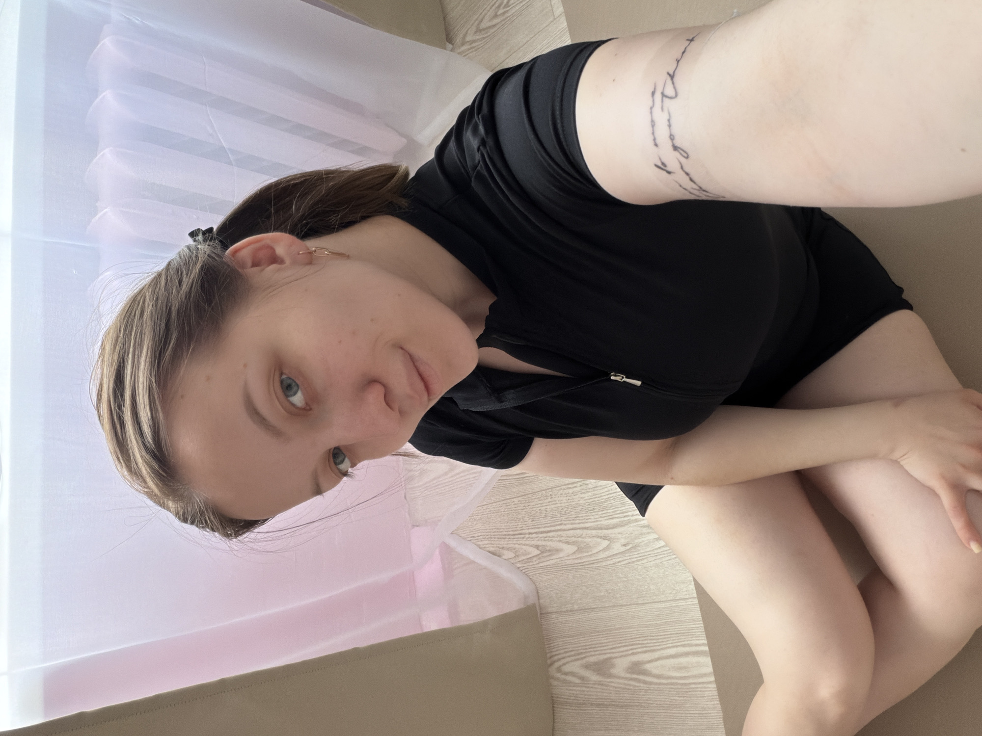 The image displays a young woman in a self-portrait, captured indoors in what appears to be a home in Reshetnikovo, Russia. She looks directly at the camera with a calm, neutral expression. Her light brown hair is styled back, and she wears a small, chain-link earring in her left ear. She is dressed in a black short-sleeved top, featuring a zipper detail, and black shorts. A script tattoo is visible on her right upper arm.
The woman is seated on a light-colored mat or rug, which rests on a light-toned wooden floor. Her legs are bent, with her knees drawn up, and one hand gently rests on her knee. In the background, a sheer, white or very light pink curtain diffuses bright natural light, indicating it is daytime. A section of light beige furniture or a sofa is also visible, contributing to the domestic setting.