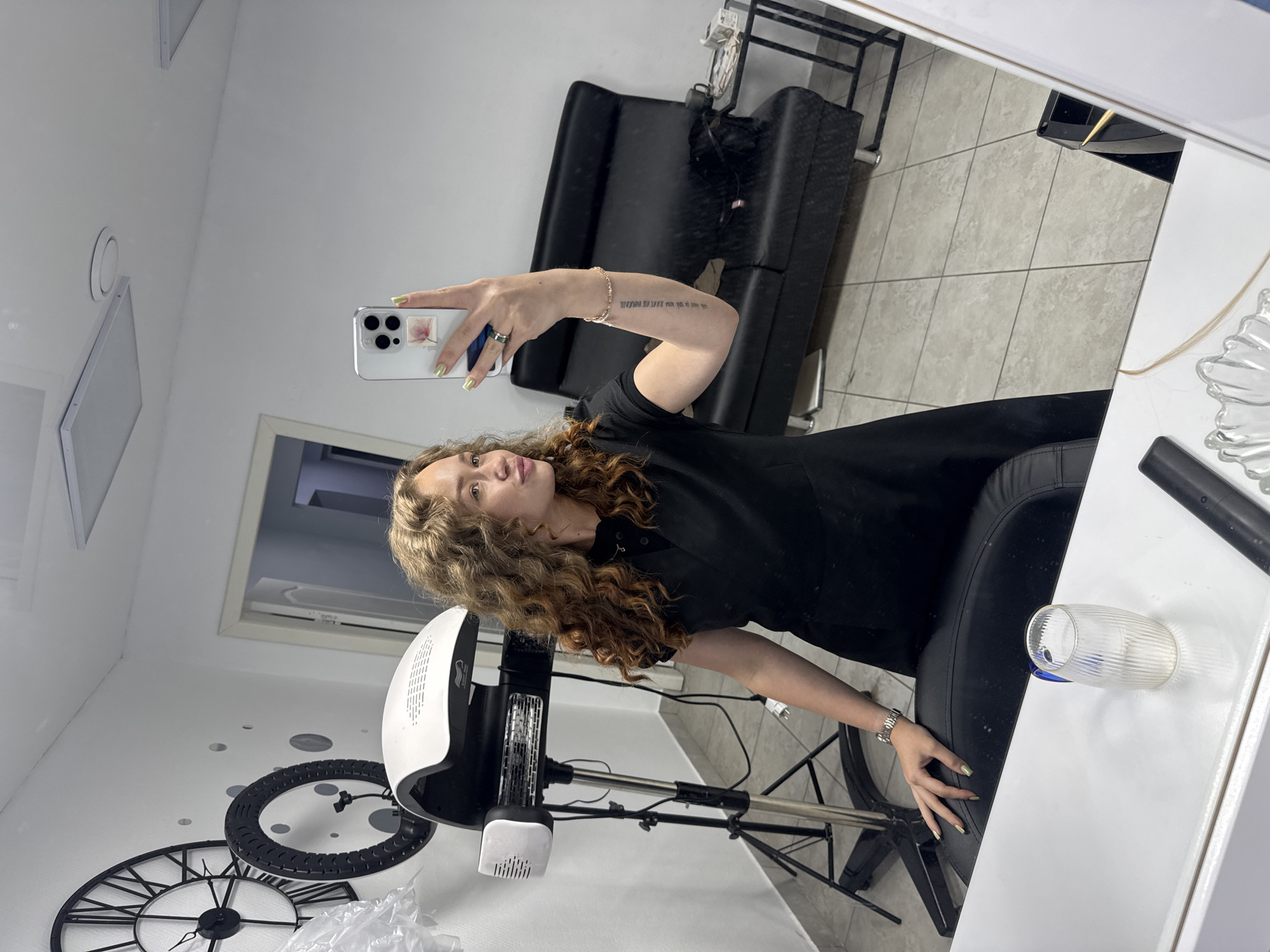 A young woman with curly blonde hair is taking a mirror selfie in what appears to be a hair salon in Chaikovsky, Russia. She is wearing a black uniform. In the foreground, a white counter and a glass of water are visible. Behind her, a professional hair dryer on a stand is positioned to her left, and a black sofa is visible to her right. A large clock face with Roman numerals is mounted on the wall behind the hair dryer. The room has tiled floors and white walls, with some ceiling lights. The overall impression is that of a clean and functional salon environment.