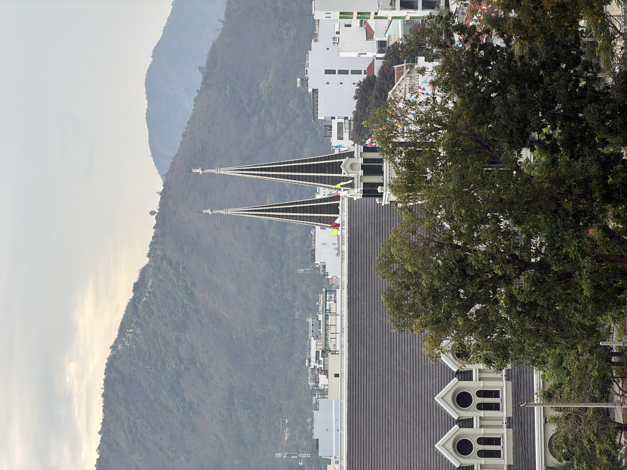 The image shows a building with two tall, pointed towers in the Bac Nha Trang Ward of Vietnam. The building has a grey tiled roof and decorative white trim around the windows and roofline. A large green tree partially obscures the building's right side. In the background, there is a cityscape with white buildings and lush green mountains in the distance. The sky is overcast.