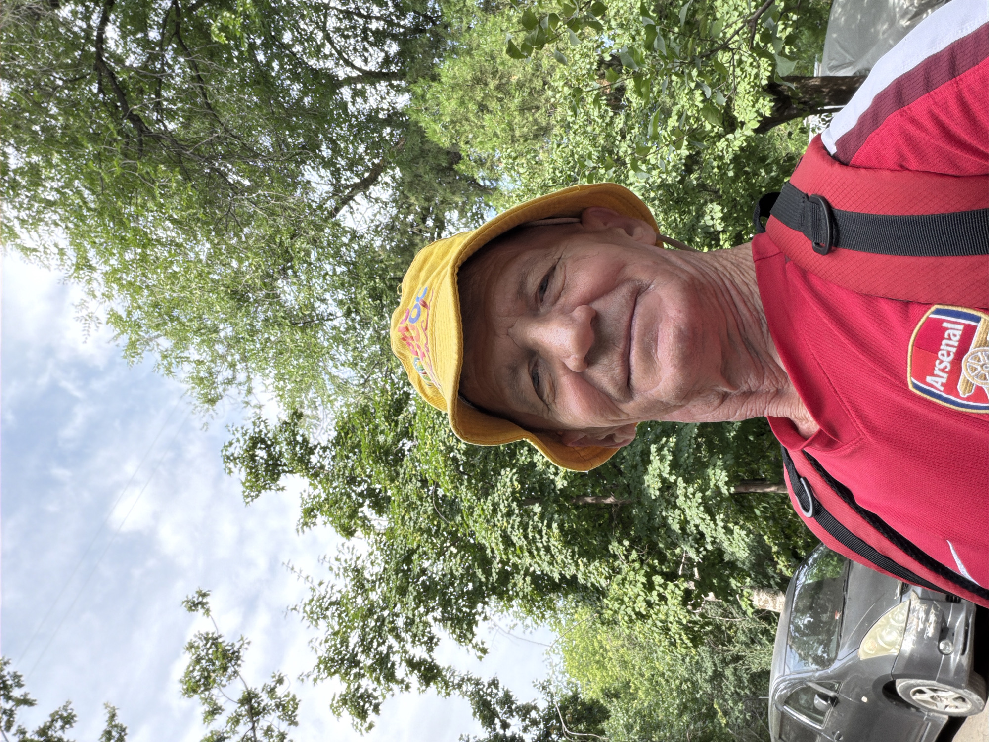 A man with a yellow bucket hat and a red shirt is standing outside in front of trees. The man is looking at the camera and smiling. He has a backpack on. A gray car is parked behind him.