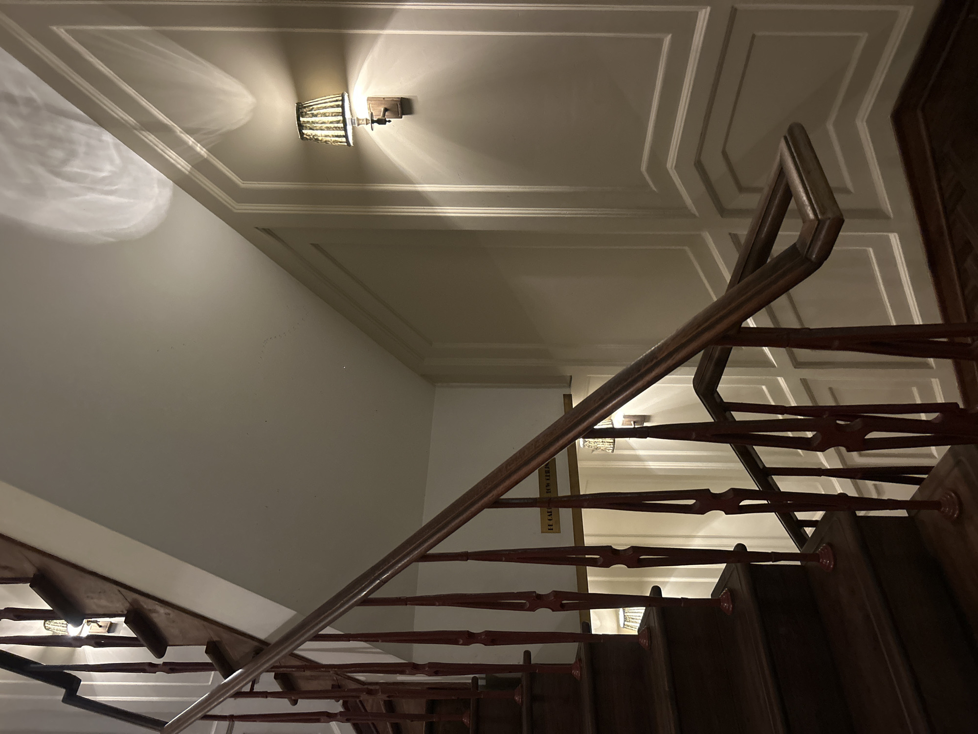 The image captures an indoor staircase with wooden steps and a railing. The walls are paneled and painted a light color. A wall-mounted lamp with a striped lampshade is visible on the right side of the image, casting light and shadows on the wall and ceiling. Another lamp is visible further down the staircase on the left. There is text on the wall behind the railing, but it is not clearly legible. The overall impression is of a well-lit, elegant interior space. The image does not contain any elements that would allow for identification of the city as Mumbai, India.