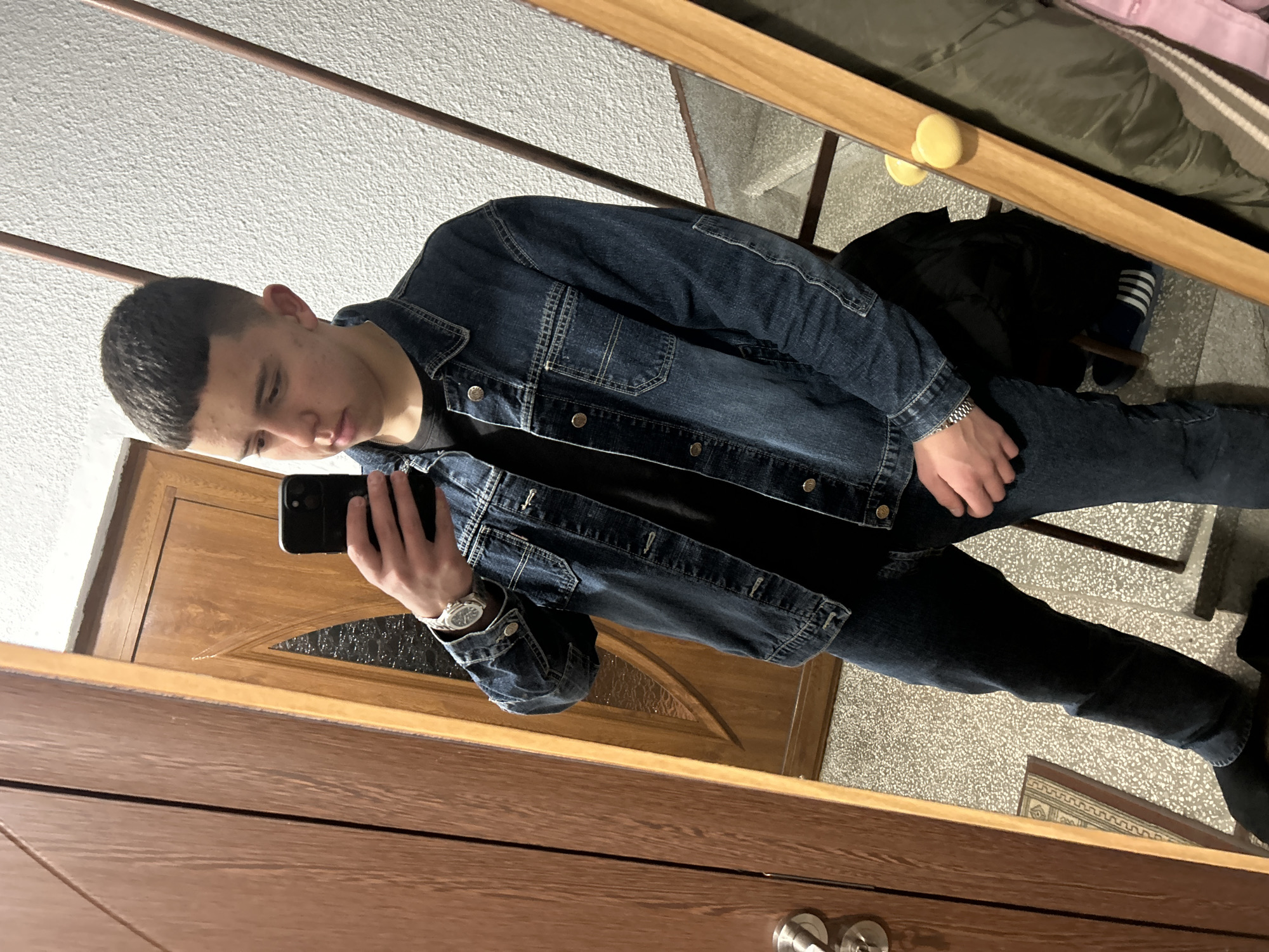 A young man is taking a selfie in a mirror indoors. He has short, dark hair and is wearing a denim jacket, a black shirt, dark jeans, and a watch on his left wrist. He holds a black smartphone in his right hand. The mirror reflects a portion of a wooden door with a decorative glass panel. The background also includes a glimpse of a room with what appears to be a coat rack and a tiled floor. Based on the presence of Zlatograd city landmarks, the image cannot be confirmed as being located in Zlatograd, Bulgaria.