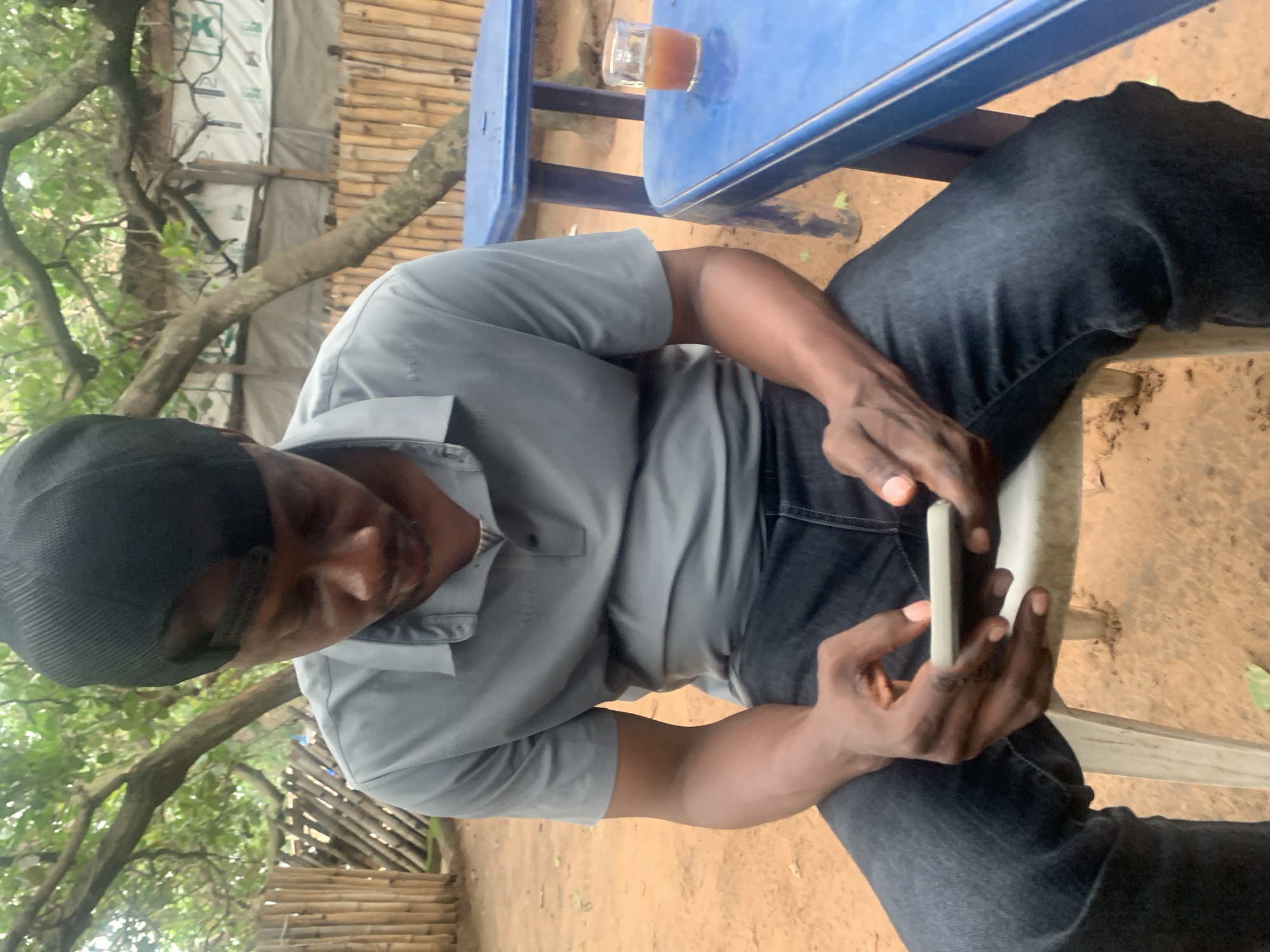 The image shows a person sitting on a chair outdoors, with a blue table in front of them. The person is wearing a grey polo shirt and dark jeans, and a black cap. They are holding a smartphone in their hands and appear to be looking at it. To the left of the person, there are trees and foliage, suggesting a natural setting. Behind them, a structure made of bamboo poles is visible, along with some white sheeting. A small glass of amber liquid sits on the blue table. The ground appears to be dirt. The lighting suggests it is daytime, and the outdoor setting implies pleasant weather. The overall scene suggests a casual moment, possibly at a cafe or a gathering place in a natural environment. There is no visible text in the image.
