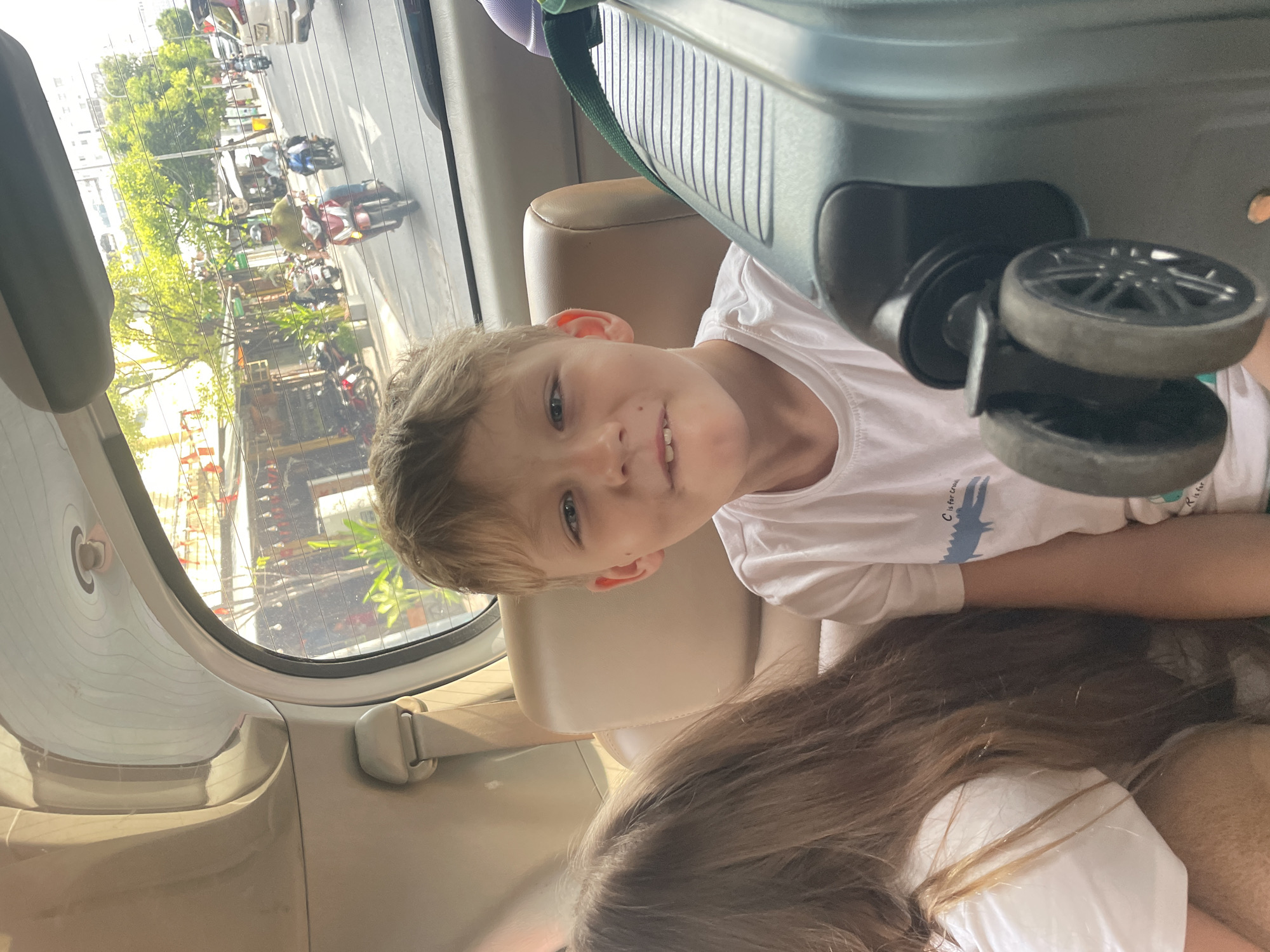 A young Caucasian boy with light brown hair and blue eyes is sitting in the back seat of a car, looking towards the camera with a slight smile. He is wearing a white t-shirt with a cartoon crocodile print and text that says "C is for cro". Beside him, to his left, is another child with long brown hair who is partially visible. The car's interior is visible, including beige leather seats and a seatbelt. Through the rear window, a busy city street in Thủ Đức, Vietnam is depicted, with buildings, trees, and numerous motorbikes and cars. The lighting suggests it is daytime, with sunlight filtering through the trees and illuminating the street scene. A grey suitcase with black wheels is positioned in the foreground of the car, to the right of the boy.