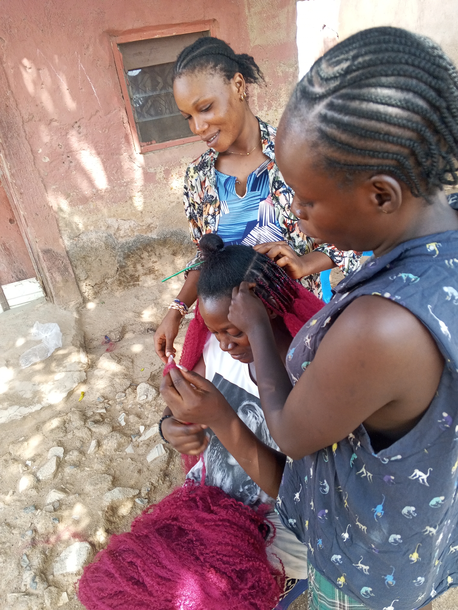 The image shows three women in an outdoor setting, likely in Jos, Nigeria. Two women are braiding the hair of the third woman. The woman being braided is seated; the braider is standing next to her. The second woman stands behind and watches the process. The braider is attaching strands of burgundy hair to the braids. A pile of burgundy hair sits near the seated woman. The area around them appears to be a dirt or sandy yard with some scattered rocks. A building with a window is visible in the background. All three individuals are African. The woman getting her hair braided is wearing a t-shirt with a grayscale image on it. The braider is wearing a blue blouse. The third woman is wearing a blue shirt and colorful outer layer.