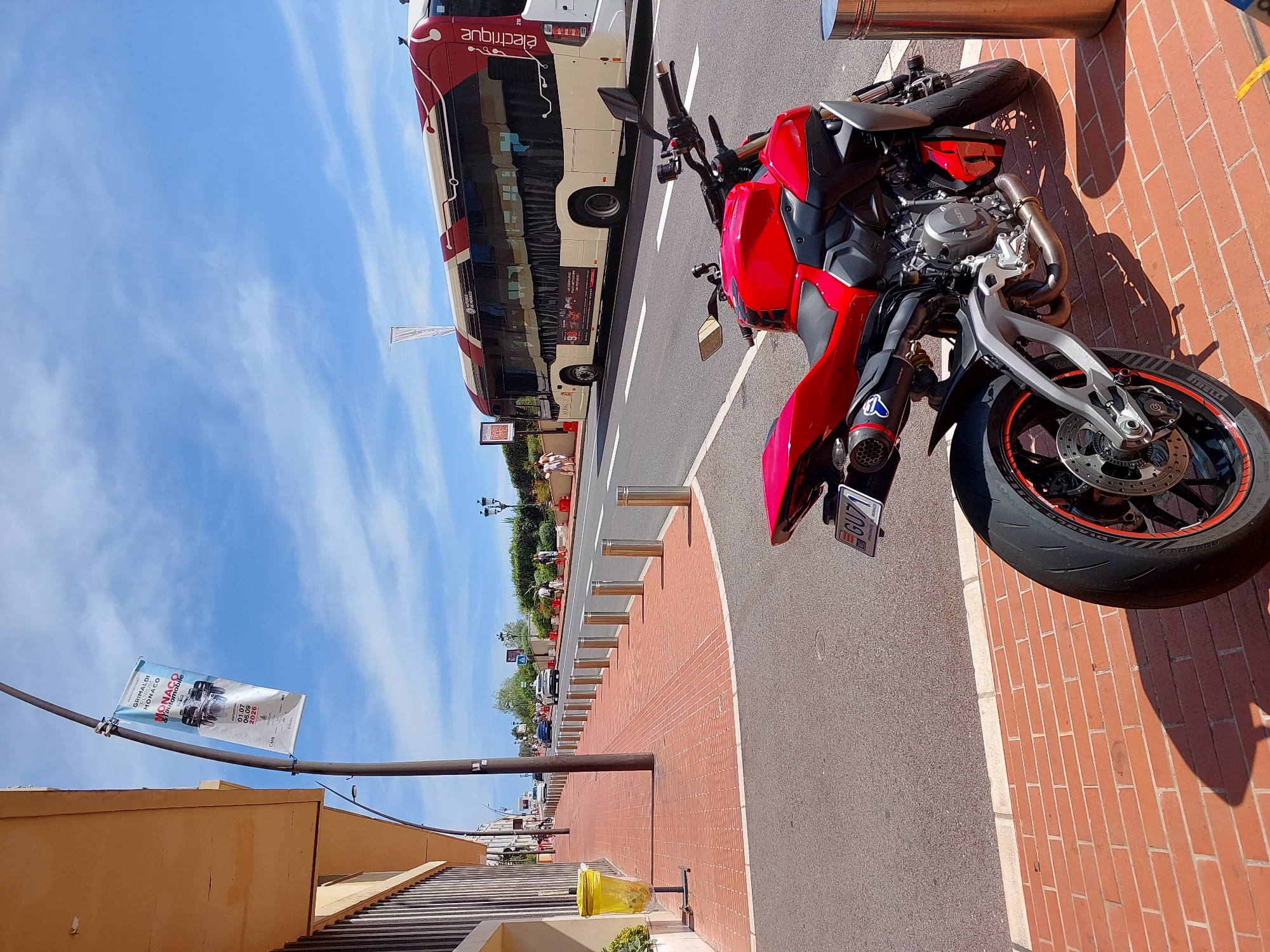 This is a street-level, eye-level, medium shot captured on a sunny day in Monaco. The foreground is dominated by a bright red, modern sport motorcycle, parked on a brick-paved sidewalk. The motorcycle is angled with its rear wheel closest to the viewer, showcasing its sporty design and exhaust. A license plate reading "GU71" is visible.
To the left of the motorcycle, a sidewalk made of red bricks leads away from the viewer, lined with metal bollards. A yellow trash can with a black stand is positioned on the sidewalk. A lamppost with a banner attached stands tall. The banner, slightly blurred, features text and an image of a race car, indicating a connection to the Monaco Grand Prix or a similar event, with dates "01.07 - 08.09 2020" visible.
Across the street, a long, modern electric bus, predominantly white and red, is driving away from the viewer. The bus has "Électrique" written on its side in stylized lettering. Several cars are visible on the road behind the bus, moving in the same direction. Further in the background, people are walking on sidewalks and there is a line of trees and greenery. The sky is blue with scattered white clouds. The overall scene depicts a lively street in Monaco, likely a tourist area, on a clear summer day.