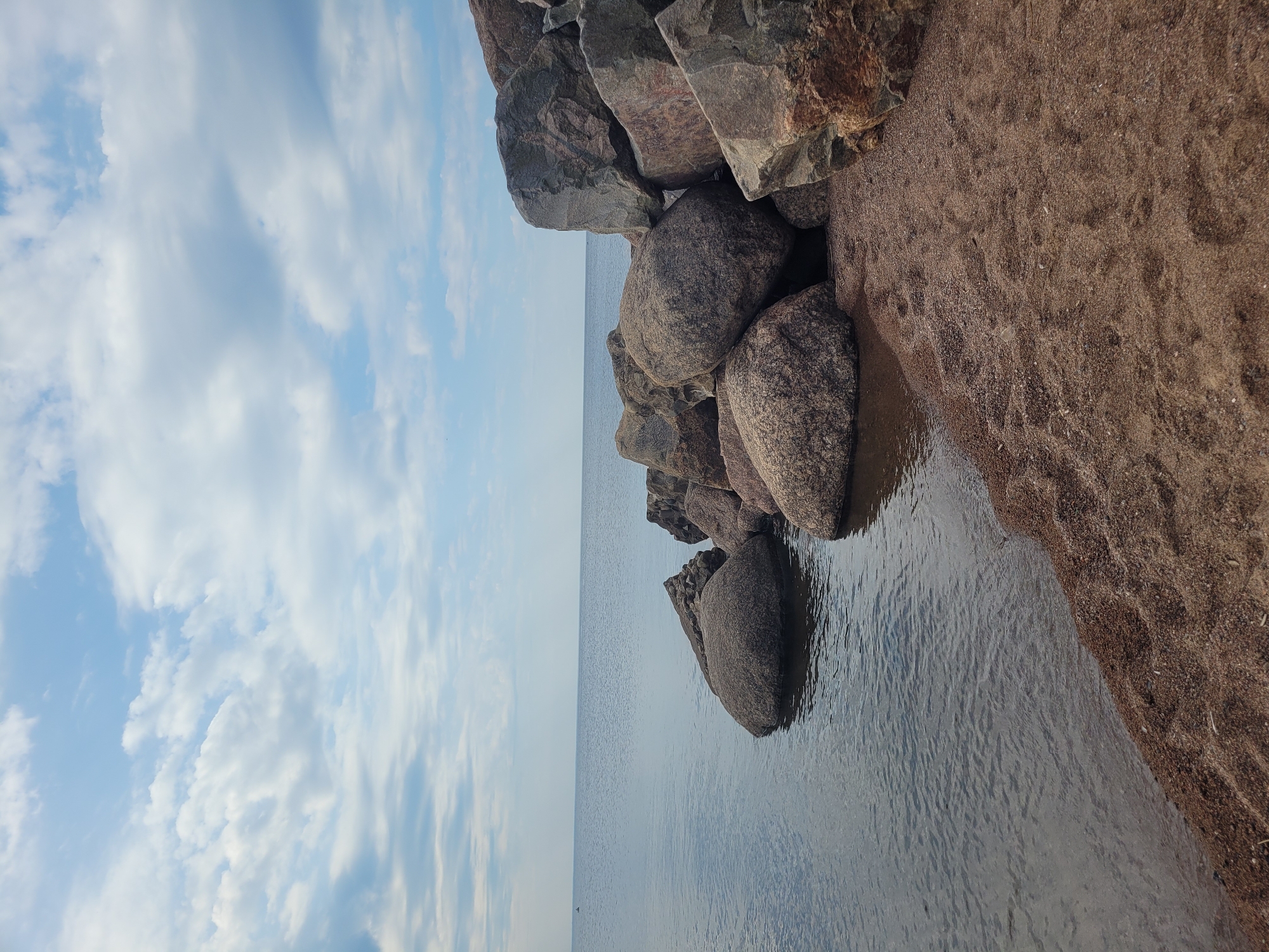 A wide shot captures a body of water and a rocky shore under a partly cloudy sky. The water is calm and appears to be a lake or sea, reflecting the pale blue of the sky. The shore is composed of large, grey boulders of various sizes, some partially submerged in the water, and a sandy beach. The sky is filled with scattered white clouds against a light blue backdrop. The overall scene is serene and natural, with no people or signs of human activity besides the arrangement of some rocks that may have been placed there. The image is taken from a low angle, emphasizing the scale of the rocks and the expanse of the water. The lighting suggests it is daytime, possibly morning or afternoon.