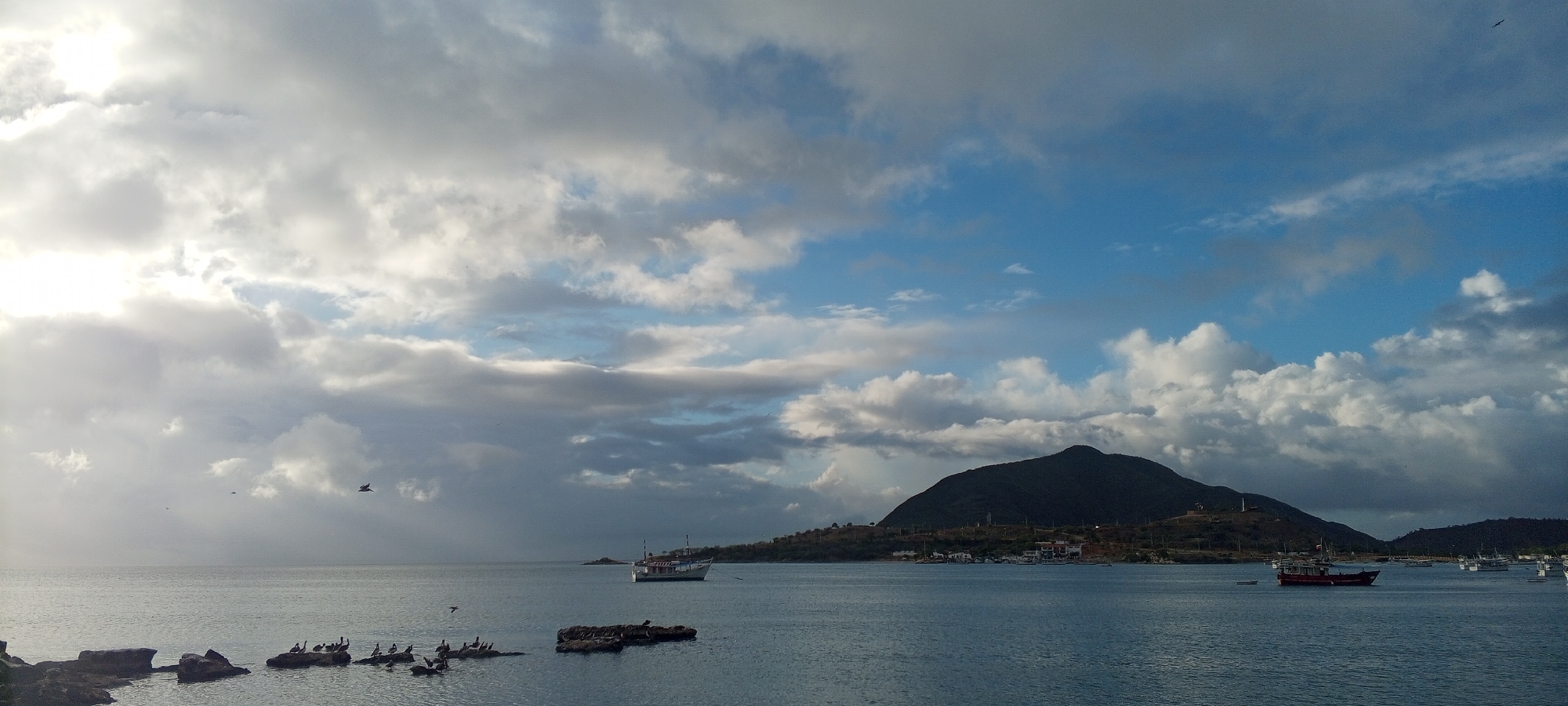 This image captures a coastal scene with a body of water, a distant hilly shoreline, and a cloudy sky. In the foreground, several dark rocks jut out from the water, with a cluster of birds perched on them. Further out, a white and blue boat is visible, and in the distance, a dark red boat is sailing. The shoreline is characterized by rolling hills covered in vegetation, with some buildings and structures dotted along the coast, particularly near the base of a prominent, dark green hill that dominates the center-right of the frame. The sky is a mix of blue patches and heavy, gray clouds, with the sun intermittently breaking through, creating areas of bright light and shadow. The overall atmosphere is somewhat dramatic due to the cloud cover. The scene appears to be a harbor or bay area, possibly near a town or village on the coast. Based on the provided context, this location is identified as Juan Griego, Venezuela.