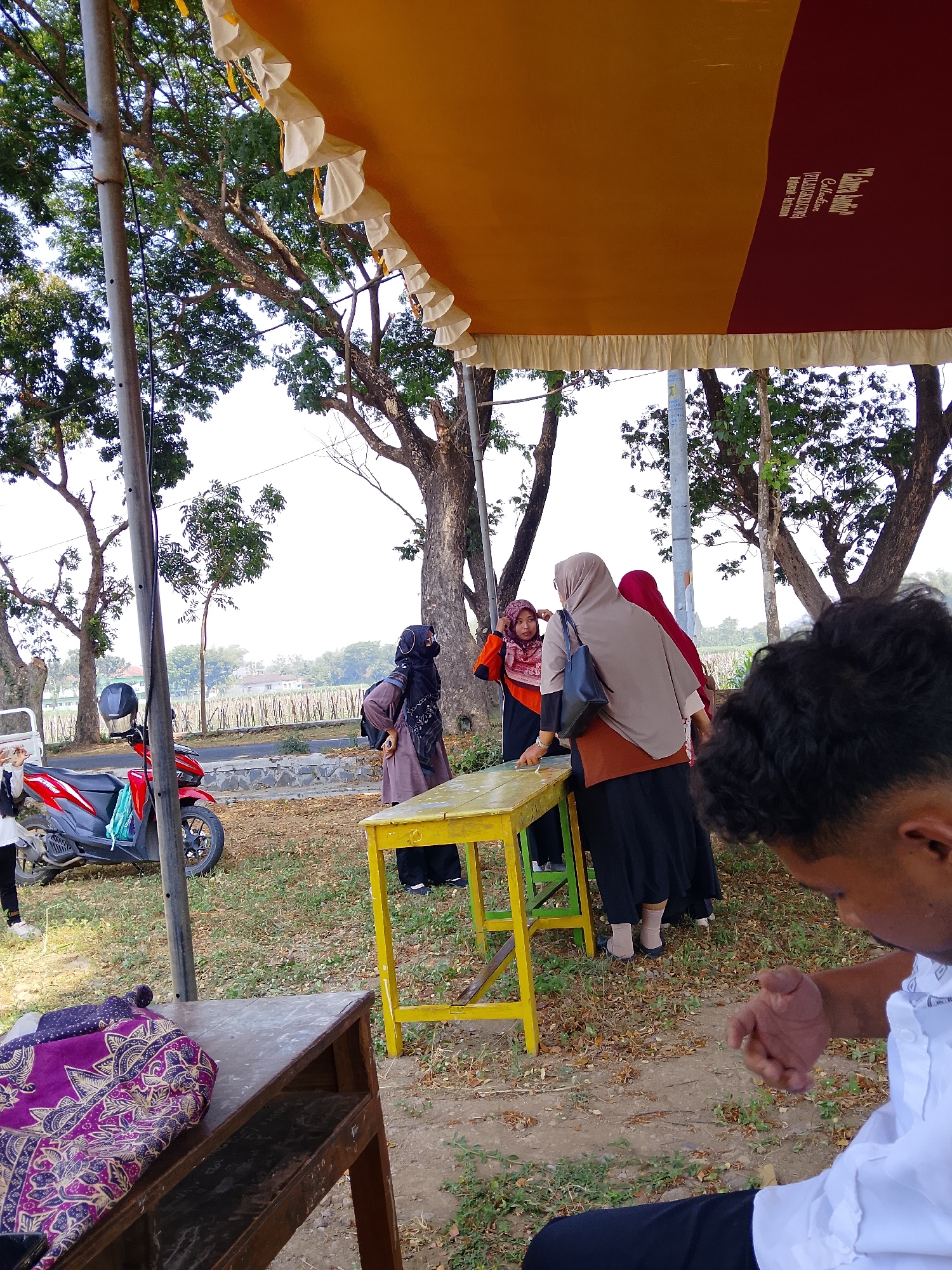 In Pelem, Indonesia, beneath a decorative yellow and burgundy tent adorned with white ruffled trim, a group of individuals are gathered outdoors. A red scooter is parked on the left side of the frame, with a black helmet resting on its seat. Several people are conversing near a yellow table. One person, facing away from the camera, is dressed in a grey hijab and a brown top, holding a black bag. Another individual, wearing a red hijab and orange top, is seen from the side. To their left, another person in a black hijab and brown attire stands with their back to the viewer. In the foreground, a man in a white collared shirt and black pants is partially visible, with his right hand extended. A wooden table with a patterned fabric is in the lower left corner. The background features trees, a road, and a field of crops under a bright sky.