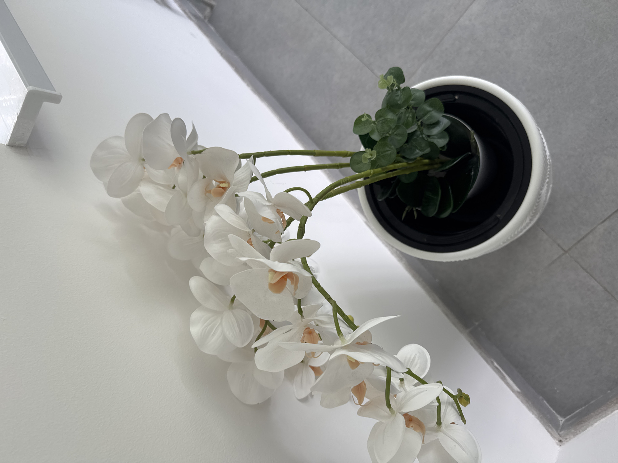 An artificial white orchid with light peach accents is displayed in a white planter. The planter sits on a gray tiled floor next to a white wall, with a white shelf visible at the top of the frame. The orchid is positioned to the left, with its delicate blooms cascading over the wall. The overall scene is clean and minimalist.