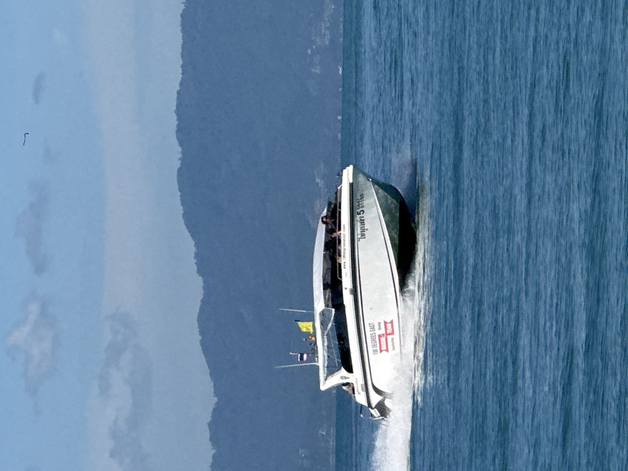 A white speed boat with a white canopy is traveling at speed on a blue ocean. The boat has "100 DEGREES EAST MARINE TEAM" written on its side in red lettering. A Thai flag and a yellow flag are visible on the boat. Several people are seated inside the boat. The boat is creating a spray of water as it moves. In the background, a chain of lush, blue-green mountains rises from the sea, with a hazy sky above. The scene is likely in the vicinity of Ko Pha Ngan, Thailand, given the textual elements on the boat and the general appearance of the landscape. A small, dark object, possibly a bird or kite, is visible in the upper right of the sky.