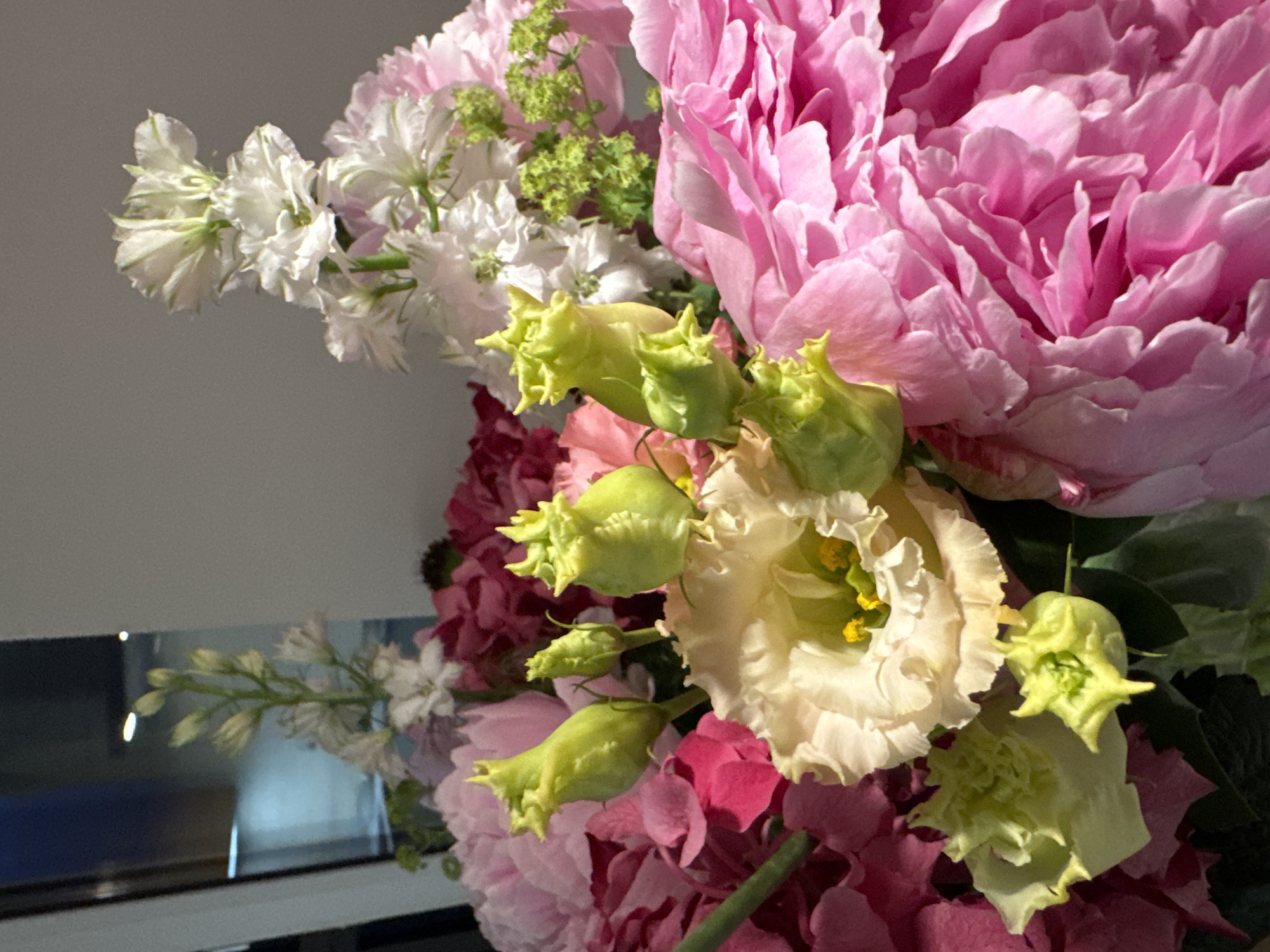 A close-up shot displays a vibrant floral arrangement, predominantly featuring large, fluffy pink peonies that occupy the foreground and right side of the frame. Interspersed among the peonies are lighter pink and deep fuchsia hydrangeas, adding depth and variation in color. Several light yellow lisianthus flowers with ruffled petals are also visible, some partially open with prominent yellow stamens. The arrangement includes a cluster of unopened green lisianthus buds with pointed tips. Tall, delicate white flowers, possibly stock or delphinium, rise from the upper left and right of the bouquet, adding height and a touch of airy texture. Small clusters of green foliage are scattered throughout. The lighting suggests an indoor setting, with soft light illuminating the flowers. The background is out of focus, showing a neutral-colored wall on the right and a dark, reflective surface with blue and white light on the left, possibly a window or a mirror reflecting a distant light source. The image does not contain any people or information indicating its location in Бараўлянскі сельскі Савет, Belarus.