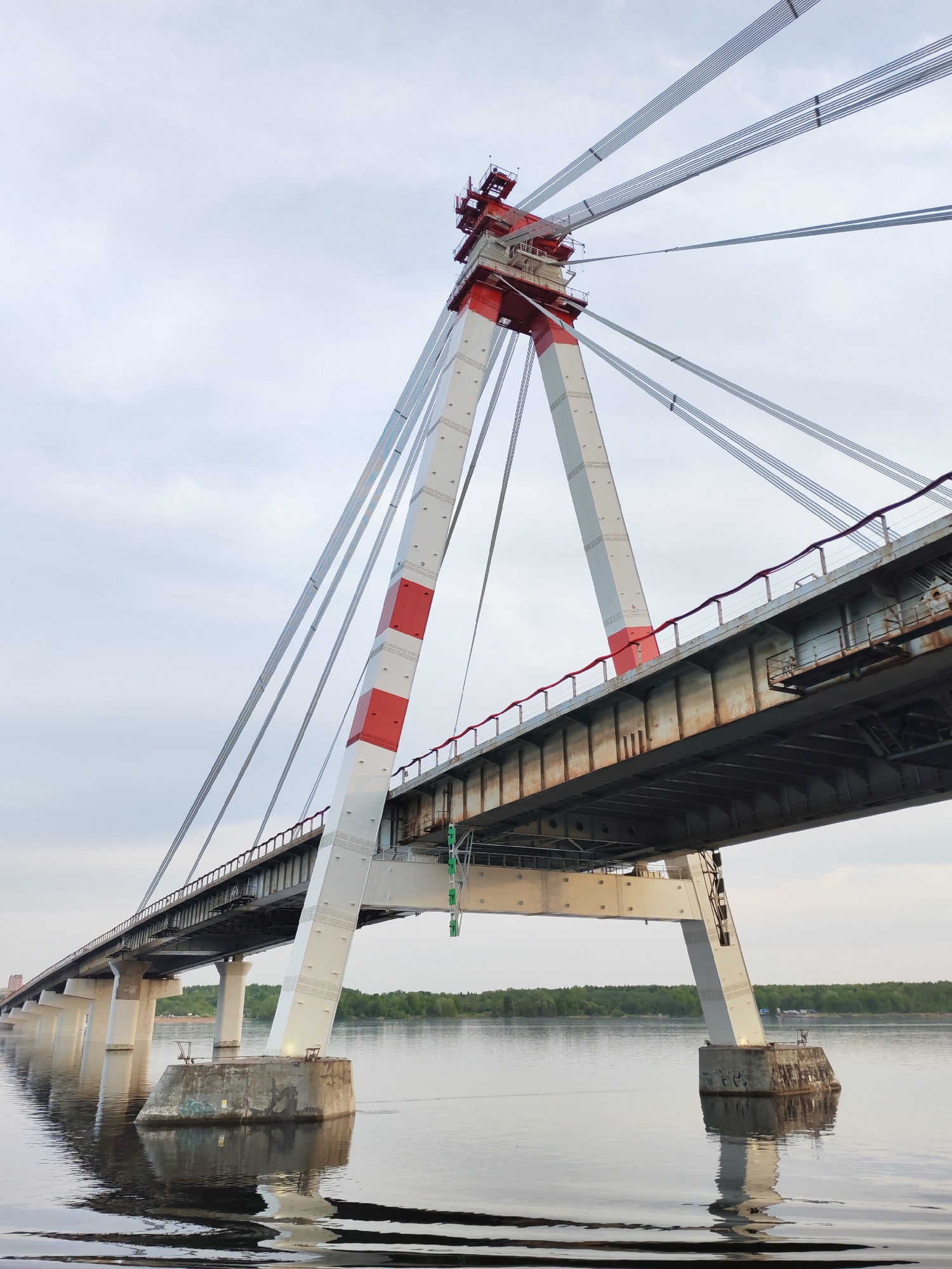 The image shows a cable-stayed bridge spanning a body of water, possibly a river or lake. The bridge's central support tower is a distinctive A-frame structure, painted white with red bands, and topped with a red structure with visible scaffolding. Numerous steel cables extend from the top of the tower to support the bridge deck. The bridge deck appears to be made of steel and has a rust-colored handrail. The bridge's supports are concrete pillars standing in the water, reflecting in the water surface. The water is calm. In the background, a line of trees is visible. The sky is overcast. The location is Unknown, Unknown.