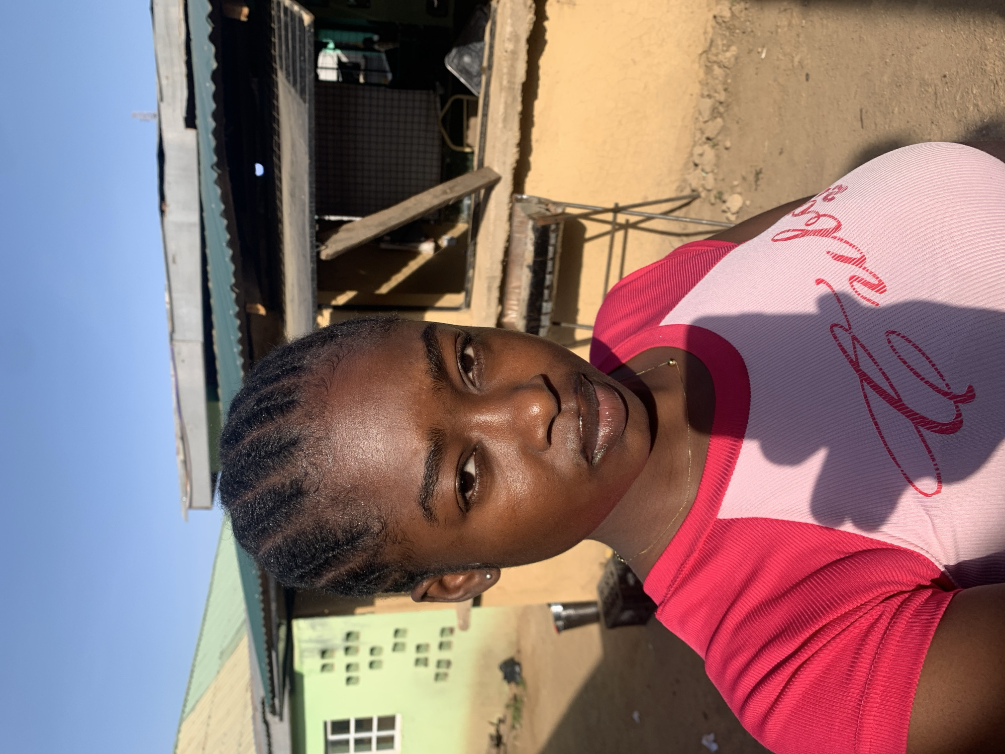 A dark-skinned woman is facing the camera. She is wearing a pink and red shirt, and her hair is styled in braids. Behind her is a building with a yellow wall and a green roof. A smaller green building is visible to the left. The background appears to be a residential area in the city of Kabong, Nigeria.