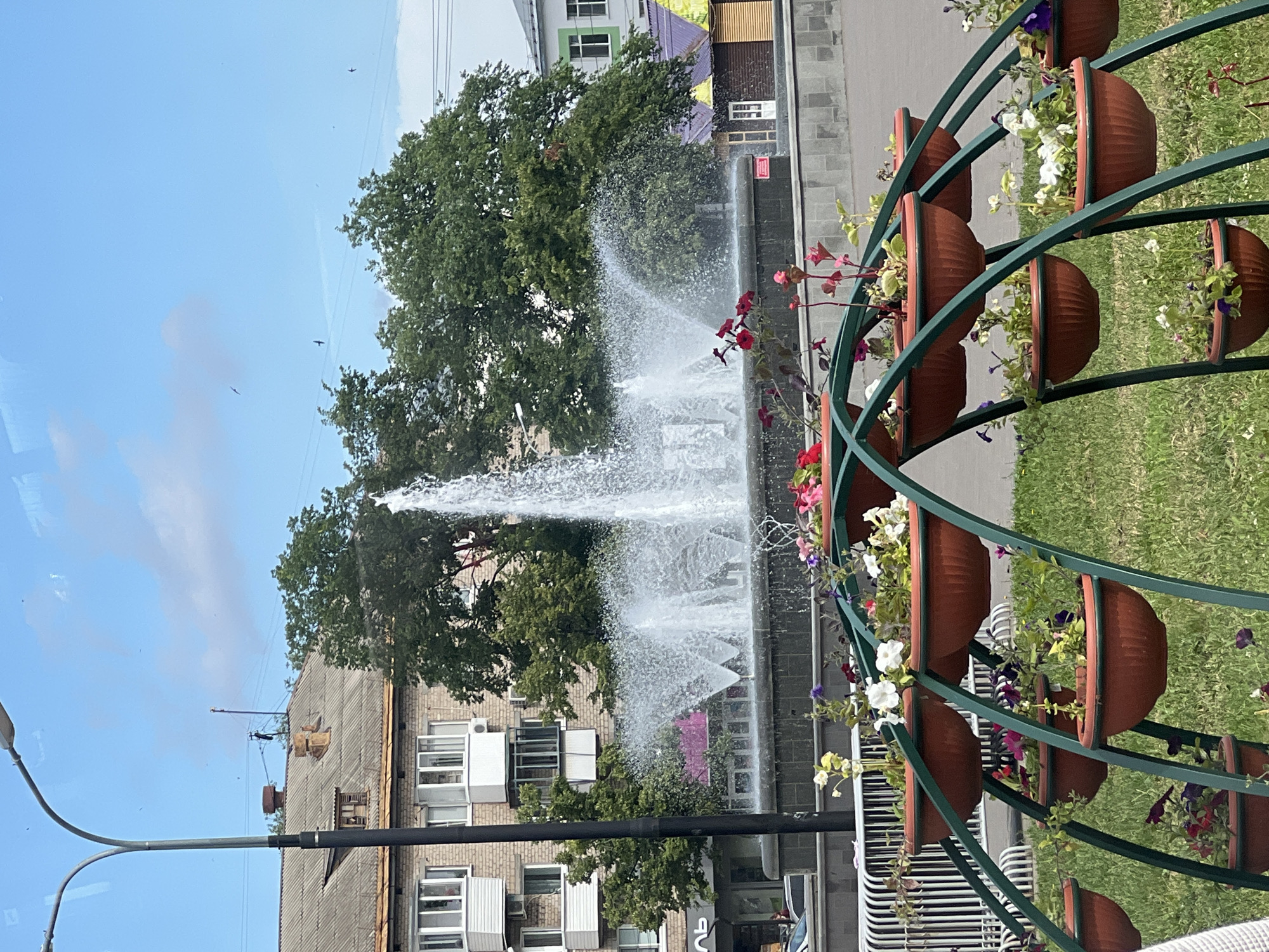 The image shows a public square in Chaikovsky, Russia, featuring a large fountain in the center surrounded by trees and buildings. In the foreground, a multi-tiered flower stand displays numerous potted plants with vibrant blossoms. The fountain is actively spraying water, creating a dynamic visual effect. The background includes a multi-story brick building, other structures, and a clear blue sky with scattered clouds and birds in flight. A lamppost is visible on the left side of the frame. The scene depicts a well-maintained urban public space.
