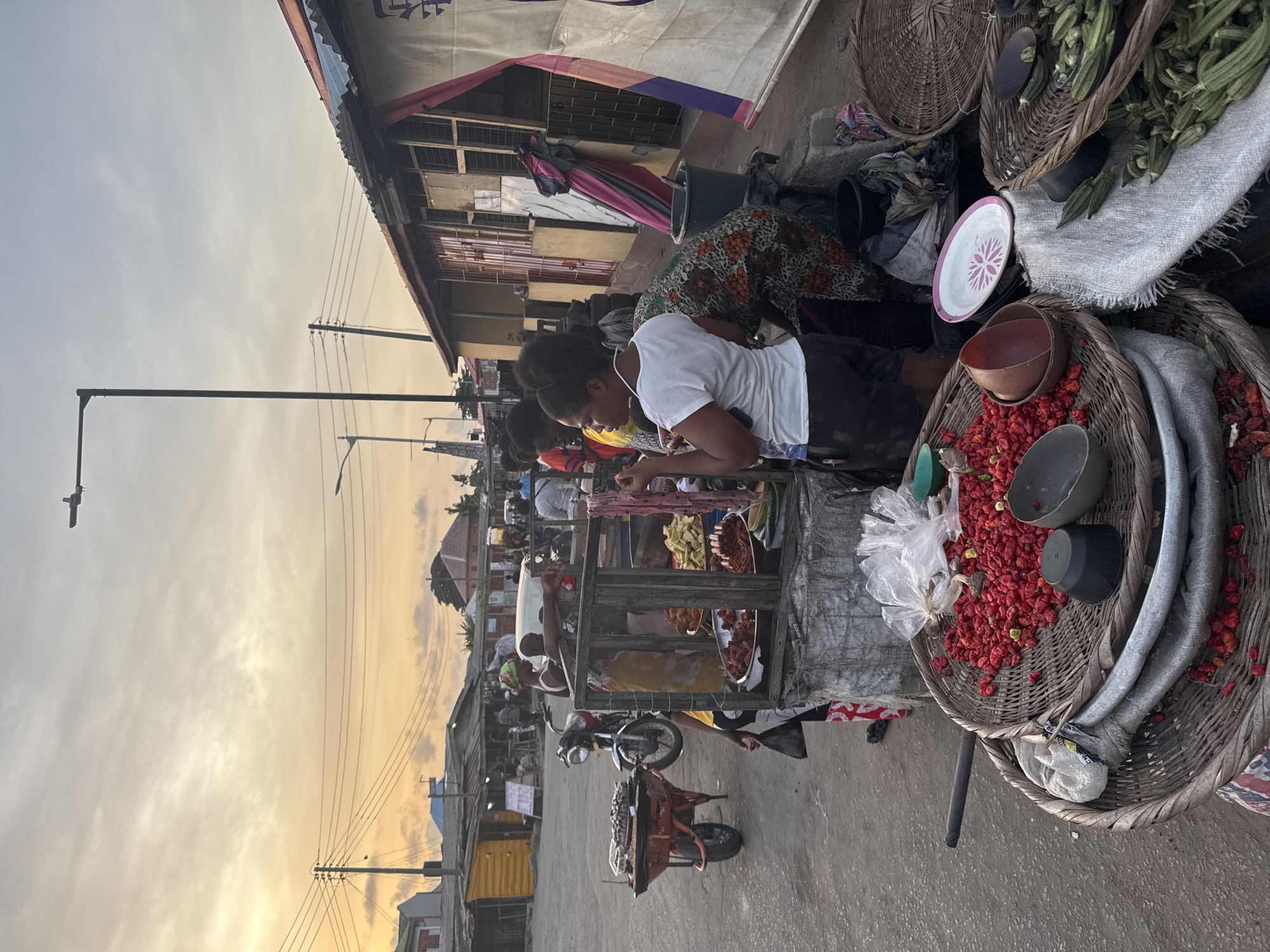 The image shows a street scene in Badagry, Nigeria. People are gathered around a street vendor's stand. The vendor has baskets of okra and red peppers. One woman wearing a white shirt appears to be a customer. The foreground is filled with produce, baskets, and bowls. A small cart and motorcycle are parked nearby. The sky is visible, and it appears to be near sunset. Buildings line the street. Overhead wires and a streetlight are also visible.