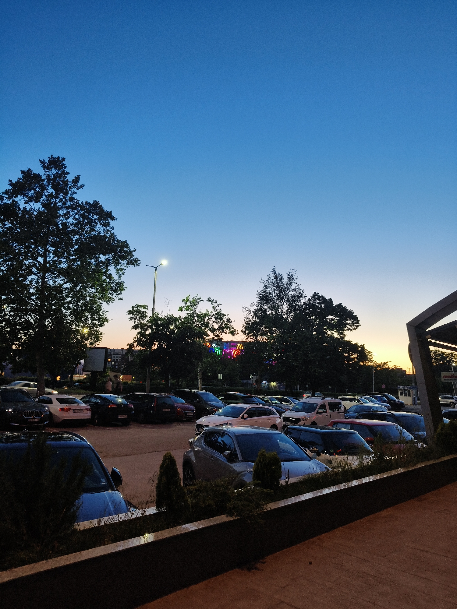 The image depicts a parking lot in Plovdiv, Bulgaria at dusk. The sky transitions from blue to orange on the horizon. Several rows of parked cars fill the foreground, with trees and a streetlight visible behind them. In the distance, colorful neon lights illuminate a building. A low wall runs along the bottom of the frame.
