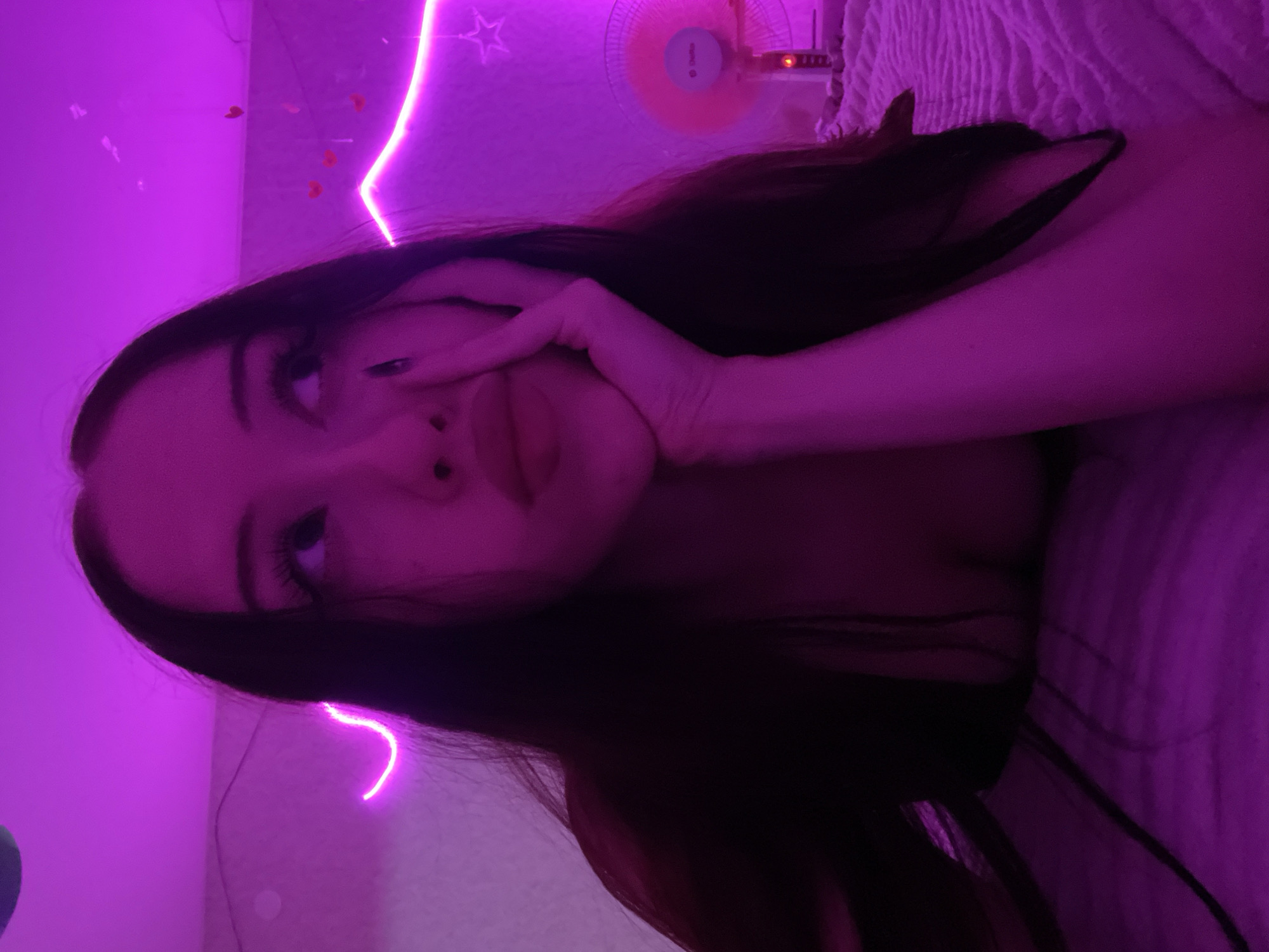 The image depicts a close-up, waist-up shot of a young woman with long, dark hair, looking upwards and to her right, with her chin resting on her hand. She is illuminated by purple lighting, which creates a vibrant and somewhat mysterious atmosphere. The background features pink neon lighting, possibly in the shape of a wavy line, and some small heart decorations on the wall. To the right, a small, pink electric fan is visible. The woman appears to be lying down on a textured surface, likely a bed or couch, also covered in purple light. The overall scene suggests a bedroom or personal space in Votkinsk, Russia, with modern decorative elements.
