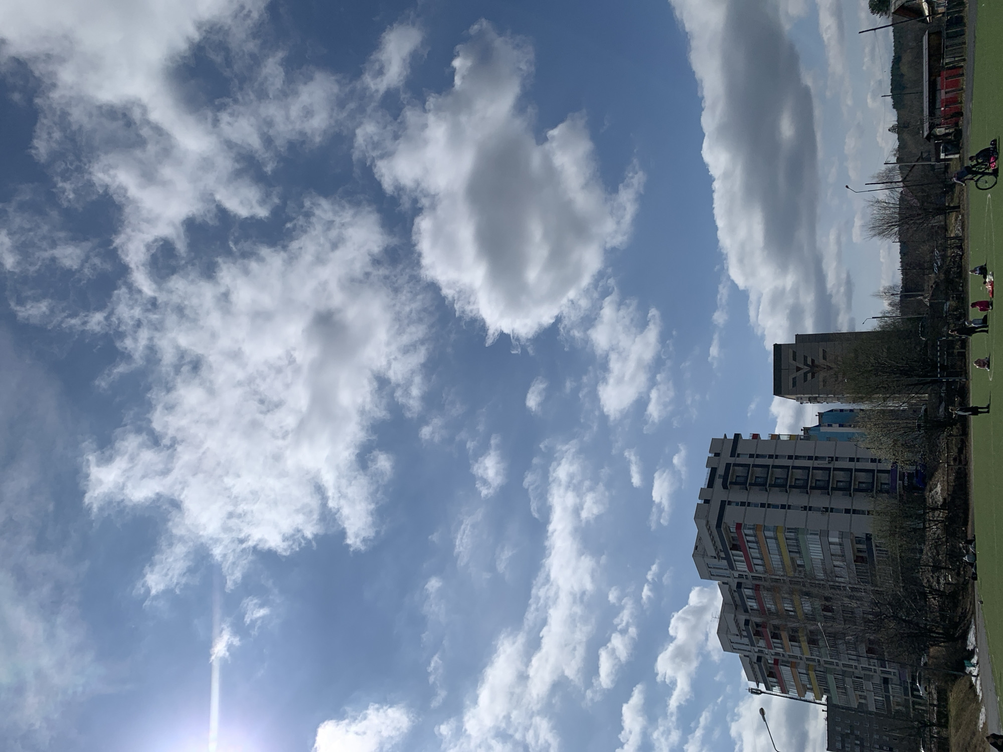 The image depicts a daytime scene in Chaikovsky, Russia, with a focus on the sky and the built environment. The sky is largely blue, filled with numerous white clouds of varying sizes and shapes. At the bottom left, there is a bright sun glare. In the lower portion of the image, there are tall residential buildings with multiple stories, the facades are mostly gray and white, with some colored accents. The buildings suggest a dense urban area. To the right of the buildings is a green area, possibly a field or park, where a few people are visible, some sitting, and one riding a bicycle. Trees with bare branches are interspersed among the buildings and the green space. The image is taken from a low angle, emphasizing the height of the buildings and the expanse of the sky. The composition is such that the sky takes up the majority of the frame.