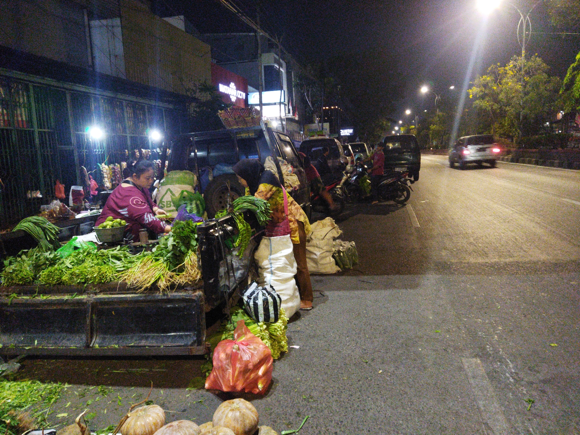 In Semarang, Indonesia, at night, a street vendor is seen by the side of a busy road, selling vegetables from a truck. The vendor, wearing a maroon shirt and a headscarf, is partially visible behind a display of fresh produce. The truck bed is loaded with an assortment of green vegetables, including leafy greens and long beans, along with pumpkins in the foreground. A woman in a maroon shirt is engaged in the transaction, while other people and motorcycles are present near the market stall. The scene is illuminated by streetlights, casting a warm glow on the area and highlighting the dynamic atmosphere of a roadside market. The road is paved, and trees line the background, indicating an urban setting.