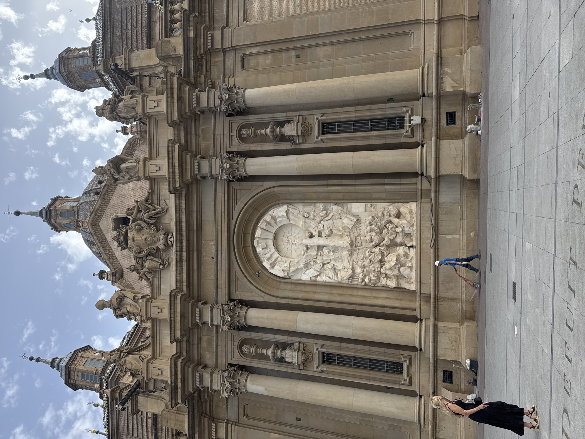 The image shows the Basilica of Our Lady of the Pillar in Zaragoza, Spain. The facade of the basilica is made of stone and features numerous architectural details, including columns, statues, and a large relief sculpture. In the foreground, a woman in a black dress and sandals stands on a paved plaza. To her right, a man in a blue shirt and a hard hat is pushing a red hand truck. The plaza is marked with large letters that appear to spell out "DEUIT PER". The sky above is partly cloudy.